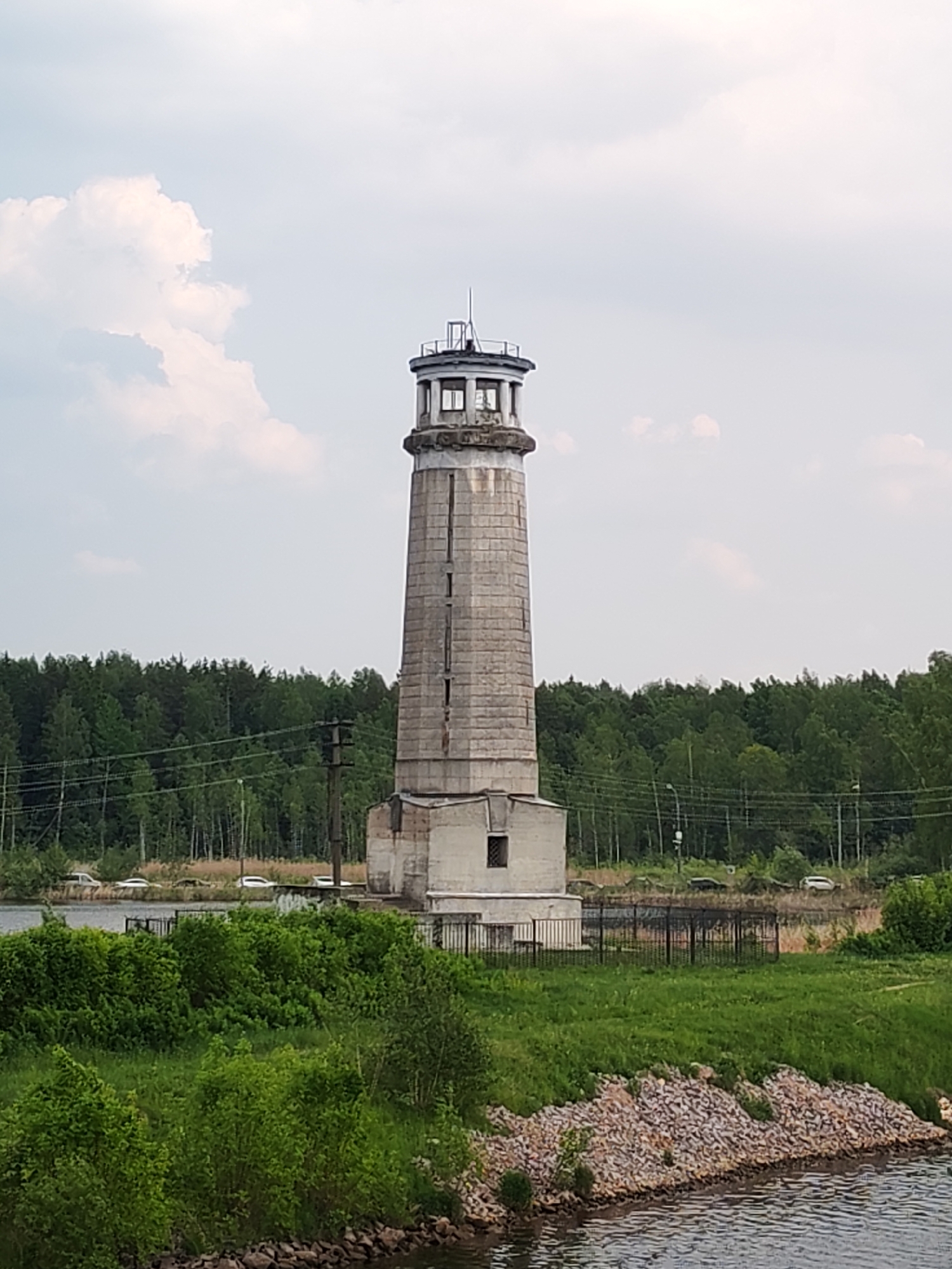 The image shows a tall, grey, brick lighthouse. The lighthouse has a circular top with what appears to be an observation deck, and a small window visible near the top. It is set on a square base. The lighthouse is located near water, with a grassy area and a line of trees in the background. The sky is overcast with some clouds. In the mid-ground, a fence separates the lighthouse from the grassy area. There are several vehicles parked behind the line of trees. The location of this structure is in the city of Unknown, Unknown.