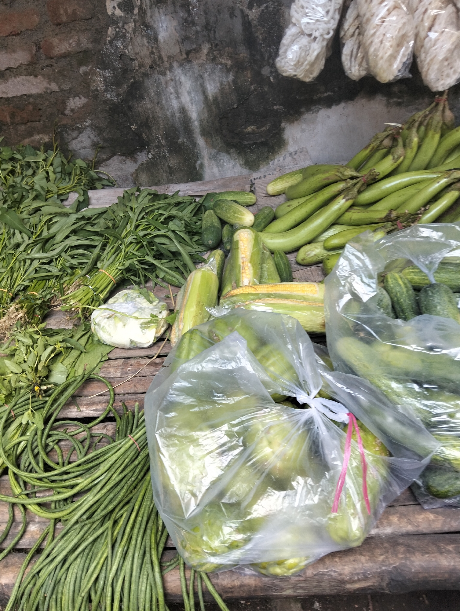A close-up, eye-level, outdoor shot displays a variety of fresh vegetables laid out on a rustic wooden surface. The vegetables include long, slender green beans, bundles of leafy green vegetables, a head of cabbage wrapped in plastic, several cucumbers, a few ears of corn, and a pile of slender green eggplants. The background consists of a weathered brick wall with patches of green moss and a gray, grimy surface. The lighting is natural and diffused. The scene appears to be a vendor's stall at a market, possibly in Muncar, Indonesia, given the context of local produce and rustic market settings often found in such areas. The composition is dense with produce, creating a textured visual of fresh food items.