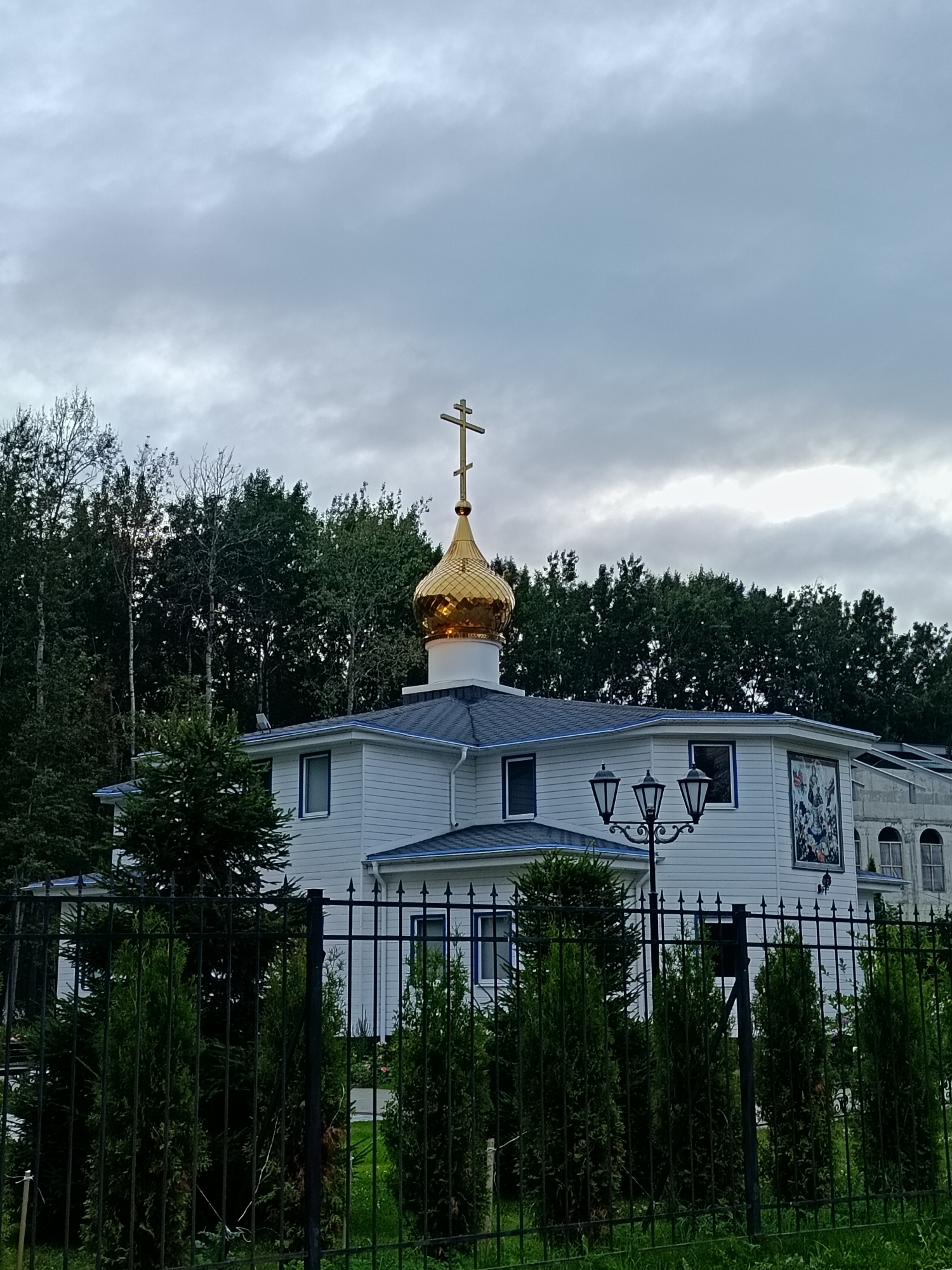 The image shows a white building with a gold dome and a cross on top, suggesting it is a church or a chapel. The building is surrounded by trees and a black metal fence. The sky is overcast with clouds. There is a lamppost in front of the building. The building has several windows and what appears to be a mural on the side. The overall scene suggests a religious institution located in a somewhat secluded, possibly suburban or semi-rural area, given the dense tree line. Without further information or context, pinpointing the exact location in Saint Petersburg, Russia, is not possible from the image alone.