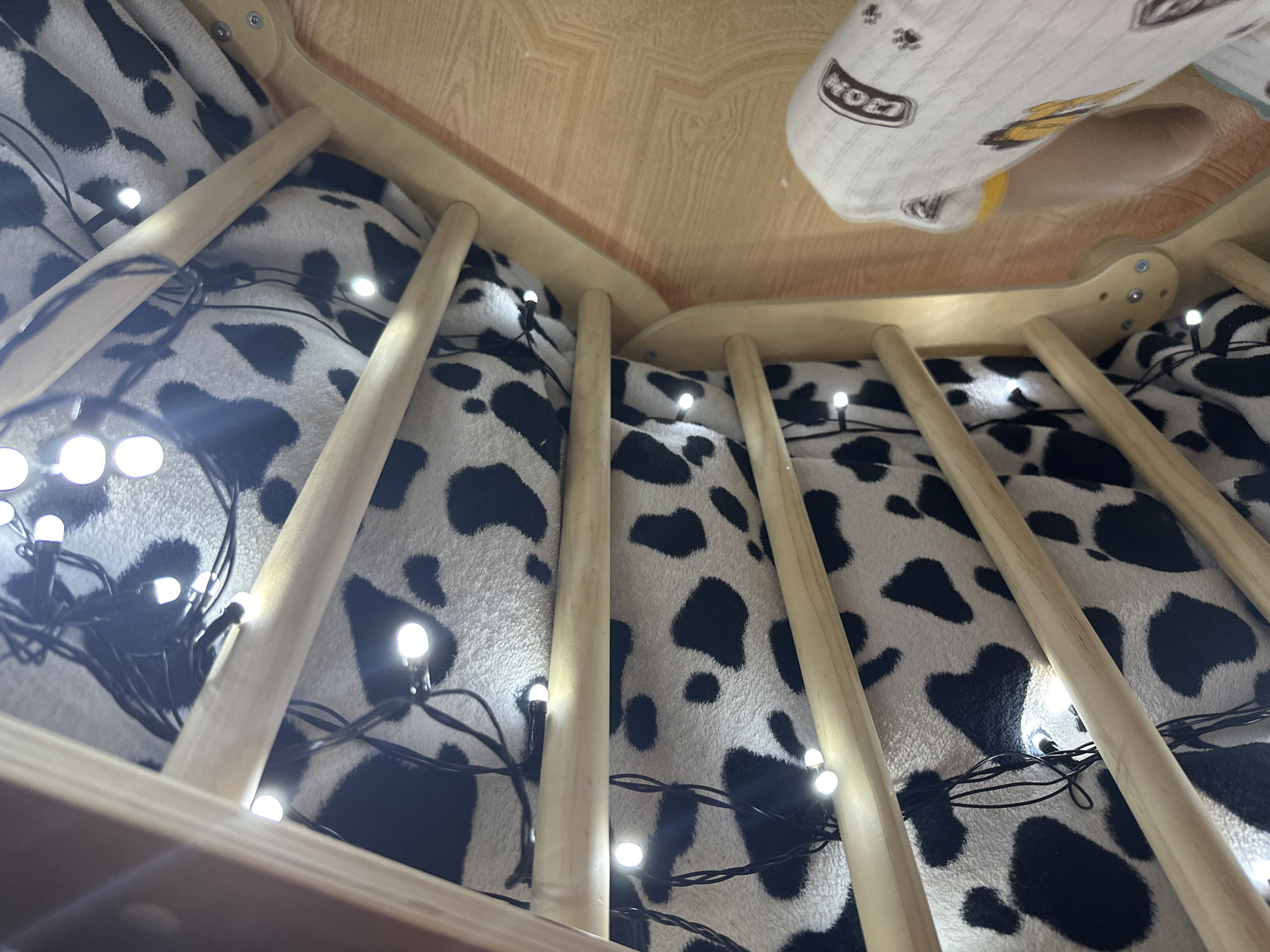 The image captures a close-up, low-angle view of a wooden structure adorned with string lights. The interior of the structure is draped with a black and white cow-print fabric. The string lights are small and circular, emitting a bright white glow, and are intertwined with the wooden supports and the fabric. The wooden elements appear to be part of a child's play structure or fort. In the foreground, a portion of a white sock with yellow and black graphics is visible, lying on a light brown wooden floor. The floor has a visible wood grain pattern. The overall scene suggests an indoor setting, possibly a child's room. The image does not contain any information that would allow for the identification of its location in Vladivostok, Russia.