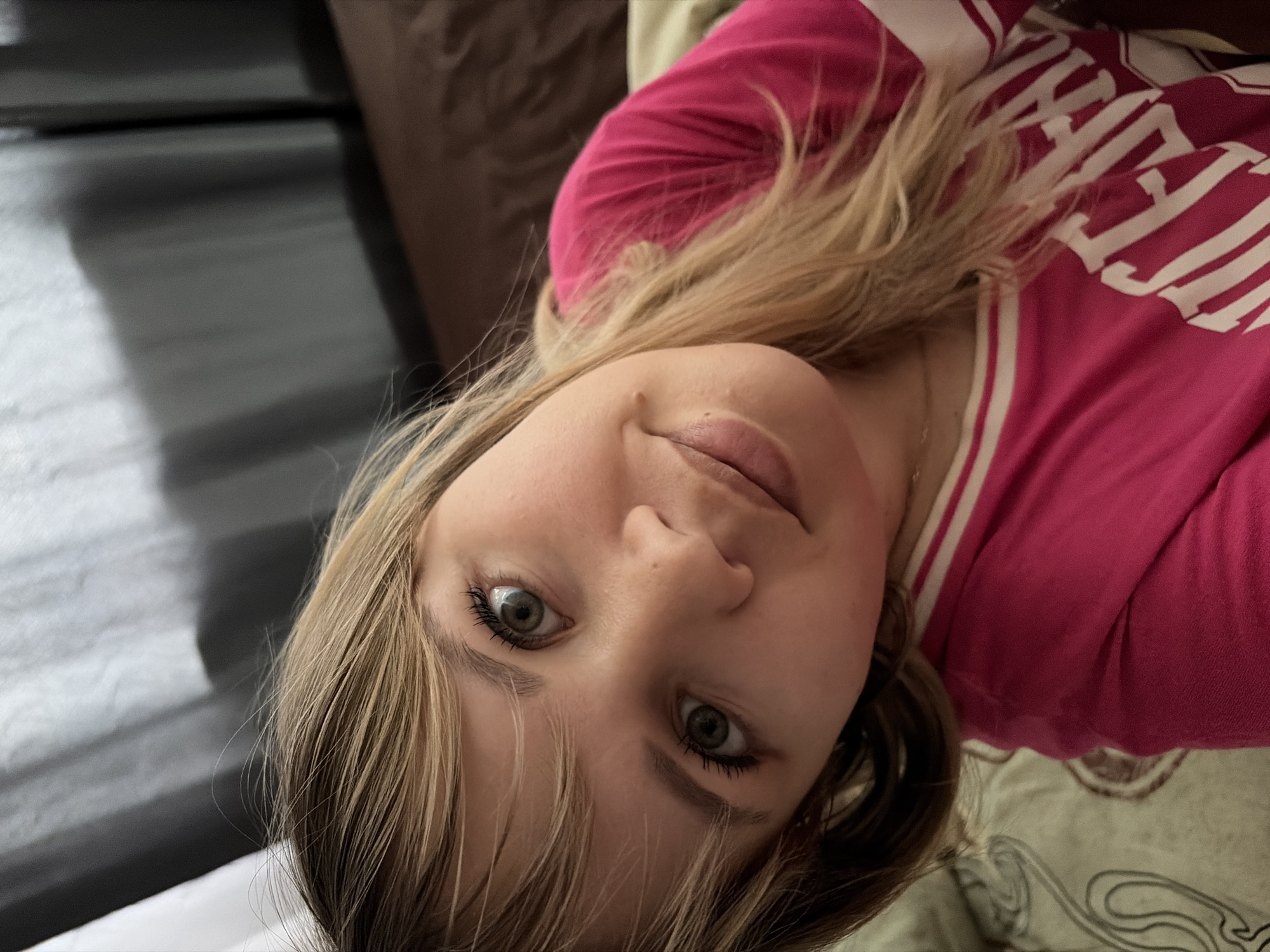 The image shows a person with light brown hair and green eyes wearing a pink shirt with white stripes. The person is indoors. The environment includes furniture with a dark wood-grain appearance and bedding. The location in the city of Chaikovsky, Russia cannot be confirmed from this image.