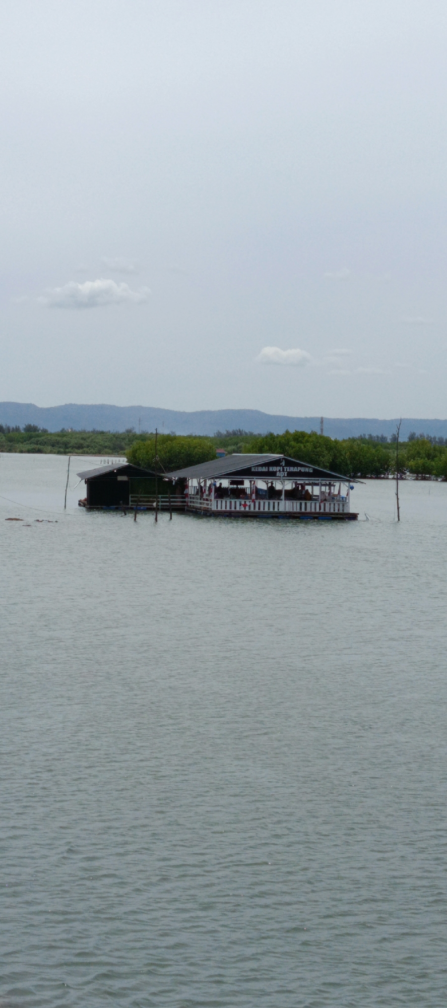 The image shows a floating restaurant located on the water in Banda Aceh, Indonesia. The restaurant is a long, narrow structure with a dark roof and white railings. Several people are visible inside the restaurant, sitting at tables. The water surrounding the restaurant is calm, with some gentle ripples. In the background, there are lush green trees and a distant mountain range under a cloudy sky. The overall scene depicts a serene and picturesque setting.