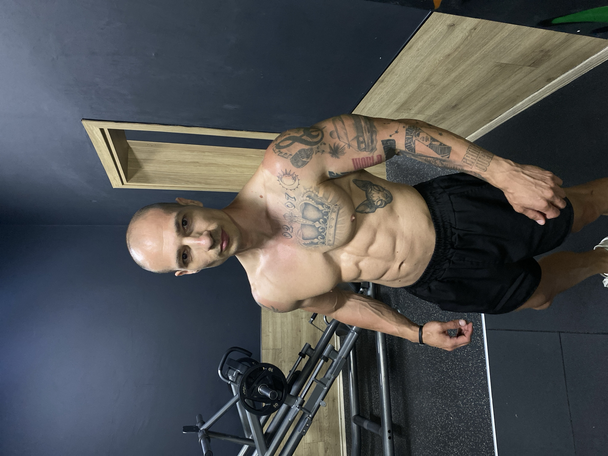 An adult male with a bald head, light facial stubble, and a neutral expression stands shirtless, directly facing the camera in an indoor gym. He possesses a muscular and lean physique, with visible abdominal definition and prominent vascularity in his arms. Numerous tattoos adorn his body, including a crown, sun, moon, and the text "02.07" on his left chest; a Sphynx cat head on his left side torso; and a full sleeve of various designs on his left arm, featuring a microphone, treble clef, car, palm trees, the number "7" with an embedded face, and the text "GAME OVER." He is wearing black athletic shorts and a plain black wristband on his left wrist.
The gym environment features dark slate-blue walls above a lower section of light wooden horizontal paneling. The floor is covered with dark grey or black rubber gym matting, separated from the wall by a thin white line. To the man's left, a grey metal leg press machine with black weight plates, marked "10," is partially visible. To his right, an open light wooden door frame is set into the wall. The lighting is even, suggesting artificial overhead illumination. This scene is located in a gym in the city of Pleven, Bulgaria.