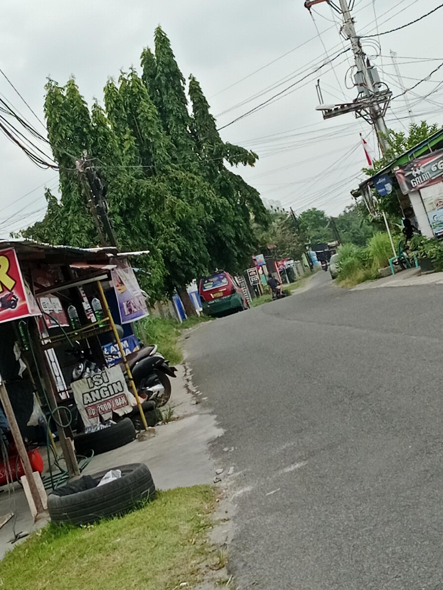 The image shows a street scene in the City of Binjai, Indonesia, on an overcast day. A narrow paved road runs diagonally through the frame, with a sidewalk and grass verge on the left. On the sidewalk, there is a small roadside stall with a tin roof, selling various items including tires, bottled drinks, and possibly engine oil. A motorcycle is parked next to the stall, and a sign in Indonesian reads "ISI ANGIN Rp 2000 / BAN", indicating an air filling service for tires at 2000 Indonesian Rupiah per tire. Further down the street, a red and green vehicle is visible, along with other roadside structures and greenery. Utility poles with numerous wires are prominent in the scene, indicating an urban or semi-urban environment. The overall impression is that of a typical street in a moderately developed area, possibly a commercial or residential neighborhood.