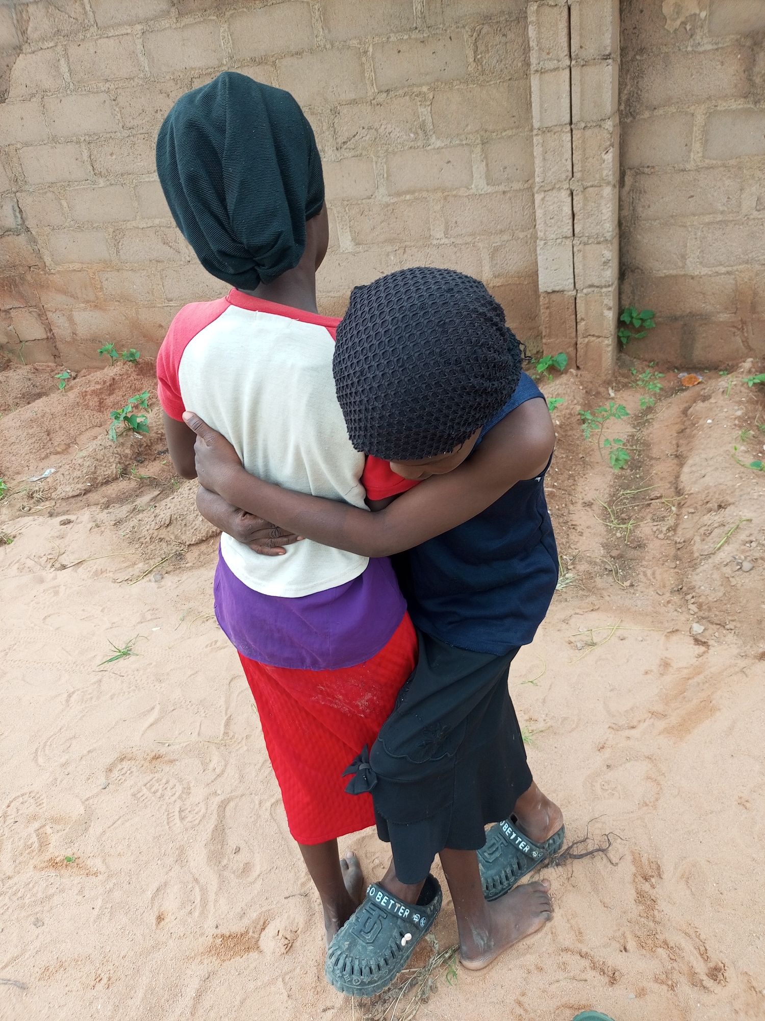 Two children, seen from behind, are embracing. The child on the left wears a black head covering, a red-sleeved shirt with a white body, a purple band around the waist, and a red skirt. The child on the right wears a black, textured head covering, a navy blue sleeveless shirt, and black pants. They are standing on sandy ground with scattered dry grass and small green plants. In the background is a light brown brick wall, with a vertical concrete pillar running down its center. This scene is located in Tempure, Nigeria.
