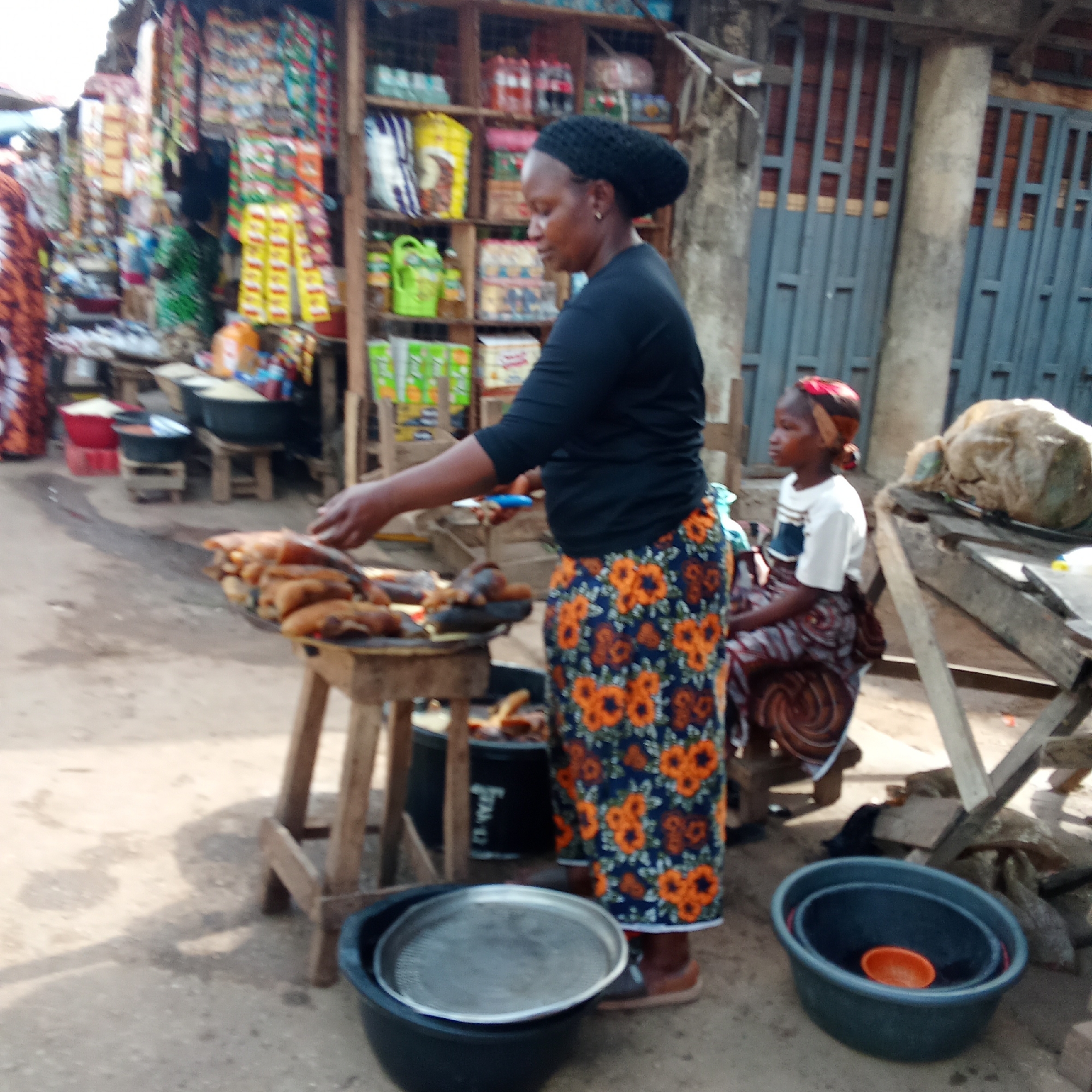 This media file captures a vibrant outdoor market scene in Akwanga, Nigeria, likely during the daytime under bright sunlight, as indicated by visible shadows on the ground.
The primary subject is an African woman standing on the left side of the frame, facing right. She wears a black long-sleeved top, a black headwrap, and a striking blue skirt adorned with orange floral patterns. Her hands are positioned over a large, flat metal tray filled with numerous pieces of what appear to be smoked or fried fish, displayed for sale on a wooden stand.
To the woman's right, a young African girl or child sits on a low wooden stool, looking towards the right. She is dressed in a white short-sleeved t-shirt and a patterned wrapper skirt, with a reddish cloth covering her head.
The background reveals a bustling market environment. Behind the standing woman, there are shelves stacked with various packaged goods, including drinks, snacks, and other provisions, indicating a general store or kiosk setup. Another person, partially visible on the far left, wears patterned clothing. Further in the background, a building or fence with grey metal gates and brickwork is visible.
At the bottom of the image, on the dusty ground, are several black plastic basins or bowls. One contains a metal sieve or tray, while another nested set includes a smaller orange bowl. These are likely used for various market tasks or for holding goods. The overall scene depicts a typical daily commerce activity in an open-air market. No legible text is visible in the image.