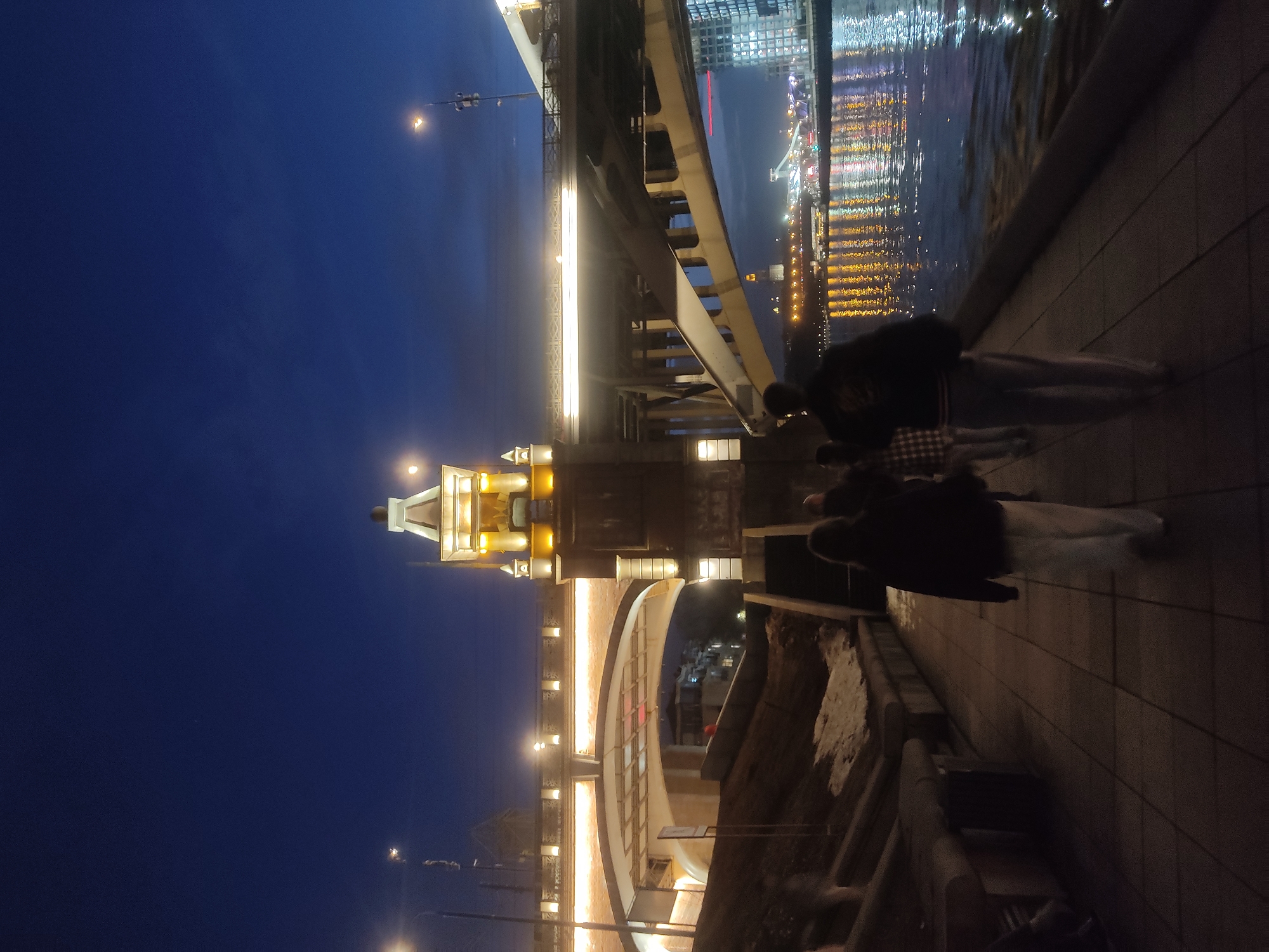 The image depicts a night scene in Moscow, Russia, specifically the Patriarshy Bridge (Patriarshiy Most). The illuminated structure of the bridge is visible, along with the reflection of city lights in the river. Several individuals are standing on the pedestrian walkway of the bridge, near the railing, seemingly observing the cityscape and river views. The night sky is a deep blue, and streetlights provide additional illumination.