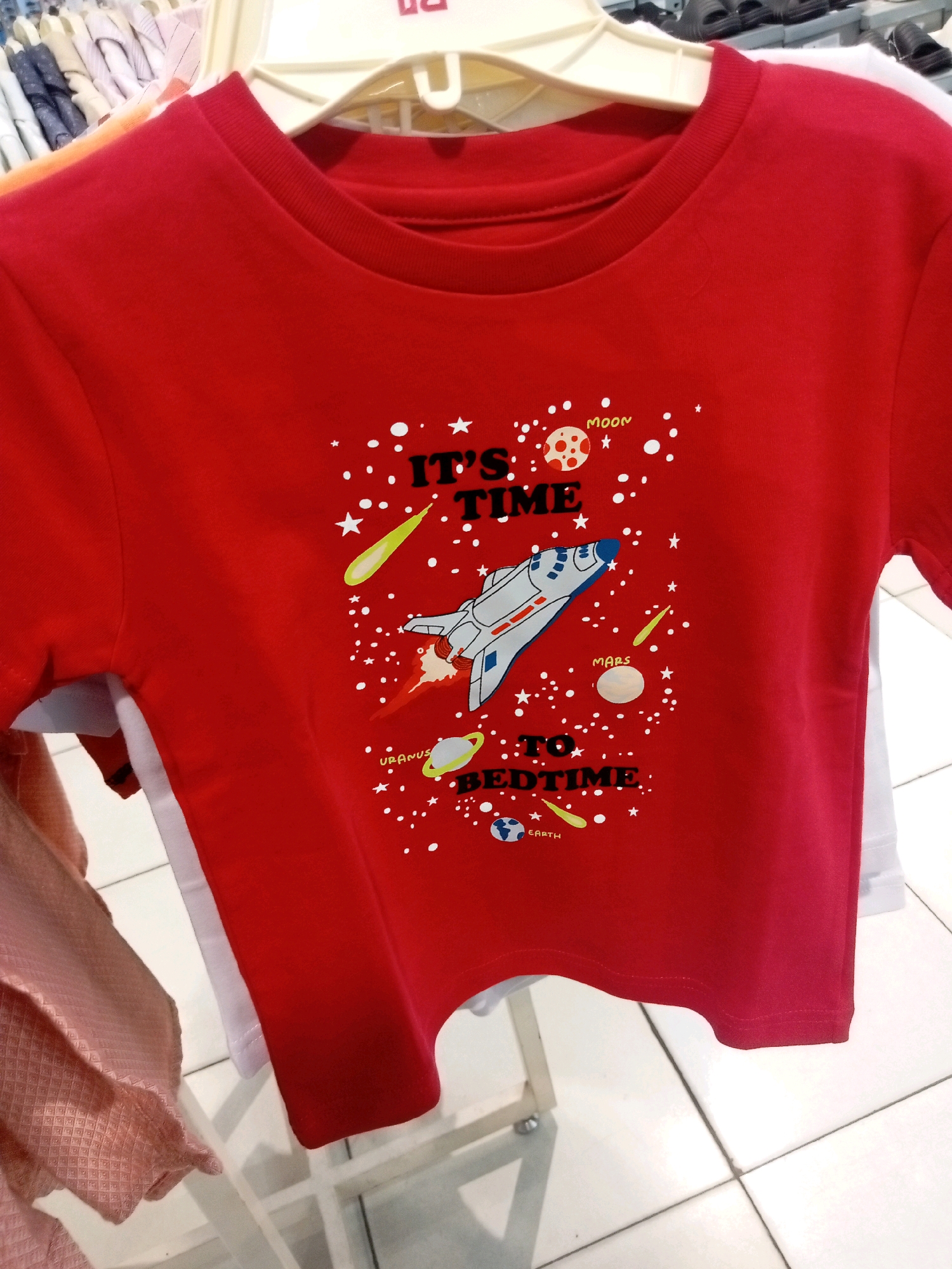 A red t-shirt with a space-themed graphic is displayed on a hanger in a retail store. The graphic features a shuttle, planets labeled "Moon," "Mars," and "Earth," stars, and comets, along with the text "IT'S TIME TO BEDTIME." The t-shirt appears to be for children. The background suggests a clothing store with racks of other garments and a tiled floor. The image was taken in Batam, Indonesia.