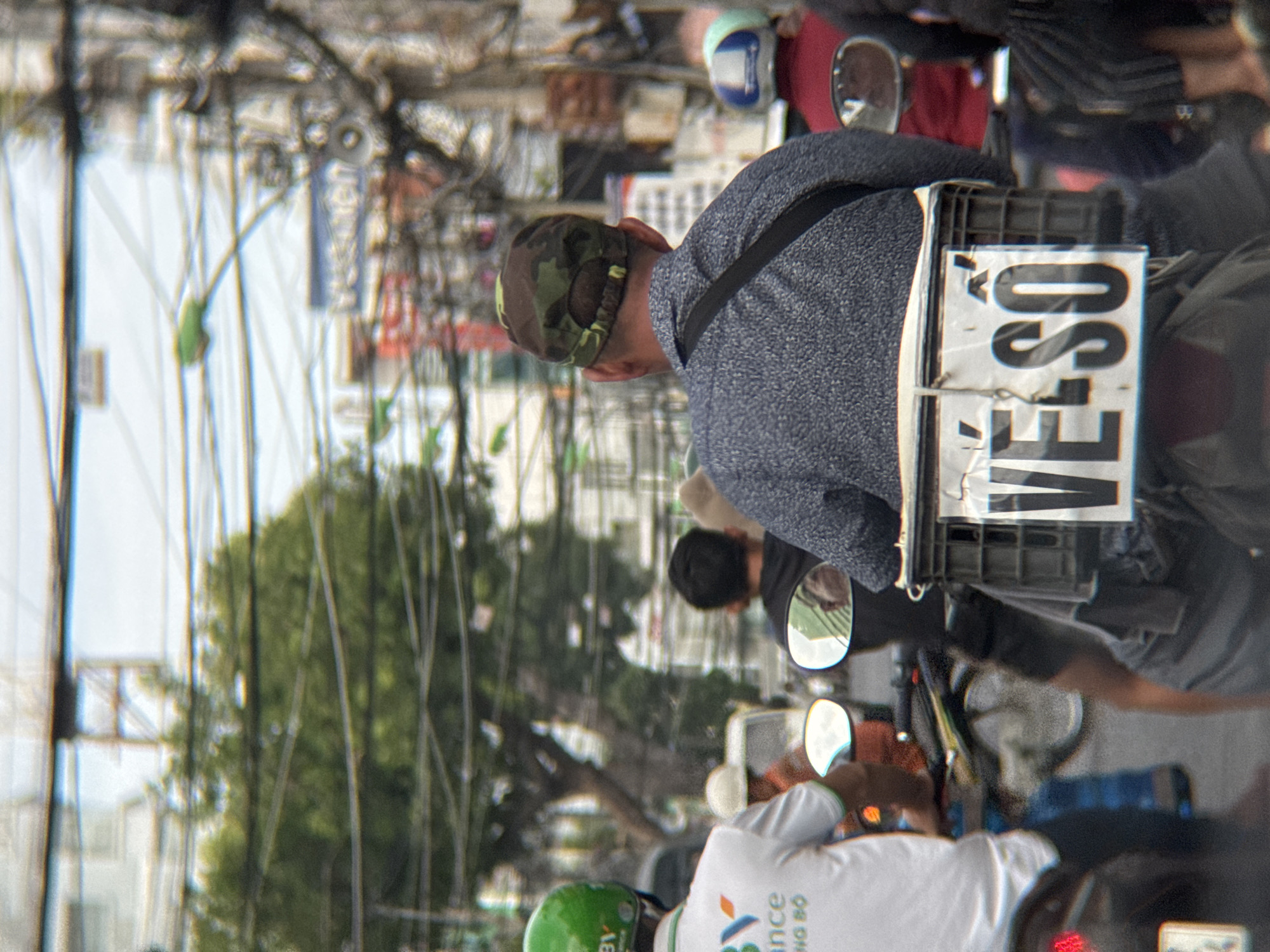 The image captures a street scene in Nha Trang, Vietnam, likely during the daytime. The foreground is dominated by the backs of several individuals on motorcycles, suggesting a traffic situation. A person in the center foreground wears a camouflage baseball cap and a textured blue-gray shirt, with a dark strap across their shoulder. To their left, another person wears a white shirt with green trim and a green helmet, with partially visible text on the shirt. To the right, a person in a red jacket is visible, also wearing a helmet.
Attached to the back of the motorcycle in the center is a black plastic crate with white signage displaying bold, dark text that appears to read "VÉ SỐ" in Vietnamese, which translates to "lottery tickets." The motorcycles and their riders are moving through a street lined with trees and numerous overhead electrical wires, a common sight in Vietnamese urban environments. Buildings and signs are visible in the blurred background, indicative of a commercial or residential area. The overall atmosphere suggests a bustling street with active transportation.