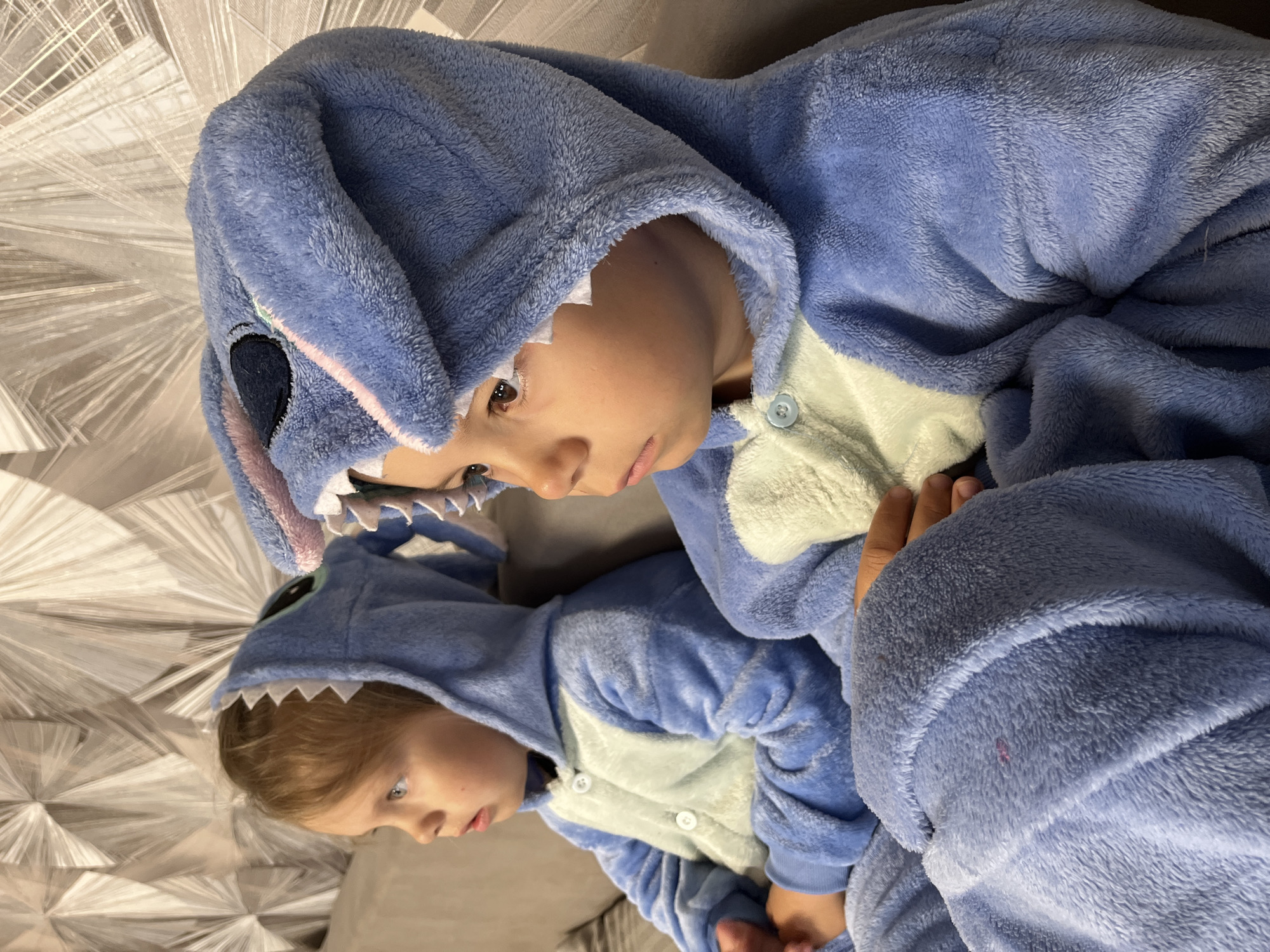 Two children are lying on a bed, wearing matching blue Stitch onesies. The location is Chaikovsky, Russia.