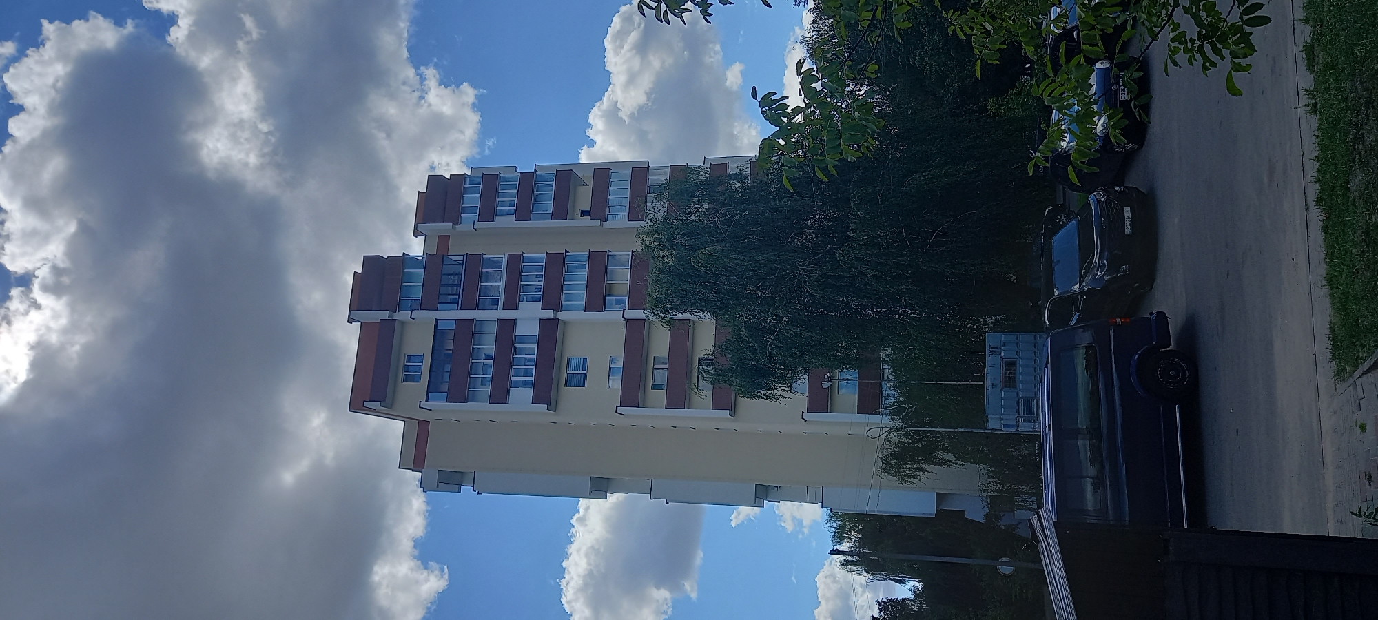 The image depicts a tall residential building in Brest, Belarus, on a cloudy day. The building is cream-colored with maroon accents around the windows and on the roofline. There are multiple stories with numerous windows and balconies visible. In front of the building, a dark blue van is parked. To the right of the van, several other cars are parked, including a black SUV with a Belarusian license plate that reads "2023MJ-1". A large, dense tree with drooping branches partially obscures the lower section of the building. Green foliage from trees and bushes frames the foreground and right side of the image. The sky is filled with both bright white and dark gray clouds, suggesting mixed weather conditions. A shipping container is visible to the left of the large tree.