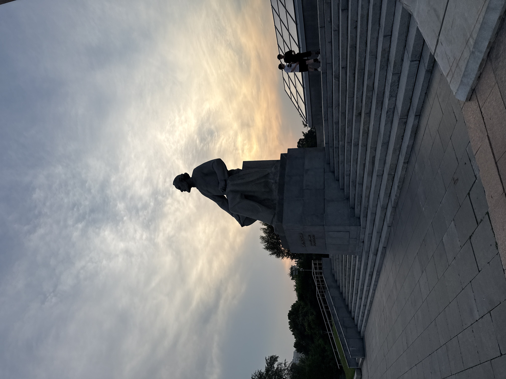 The image shows a large, stone statue of a seated man, silhouetted against a cloudy sky with hints of sunset colors. The statue is on a tiered stone pedestal, which is part of a grand staircase. Two people, one wearing a white cap and the other a black cap, are standing on the stairs, looking towards the statue. To the right of the stairs, there is a modern building with a glass roof, also silhouetted. The scene appears to be outdoors in a park or public square. Based on the inscriptions visible on the pedestal of the statue, it is identified as the monument to Maxim Gorky, located at Krymskaya Embankment in Moscow, Russia. The inscription on the statue's pedestal reads "МАКСИМ ГОРЬКИЙ" (Maxim Gorky). The time of day is likely late afternoon or early evening, given the lighting conditions. The environment is characterized by stone architecture, greenery in the background, and a dramatic sky.
