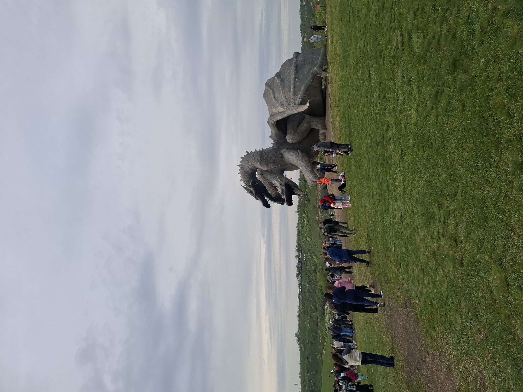 A large stone dragon sculpture is the focal point of a scene in Алисово, Russia. The sculpture is positioned on a grassy field where a large group of people are gathered along a path in the field. The sky is overcast with clouds.