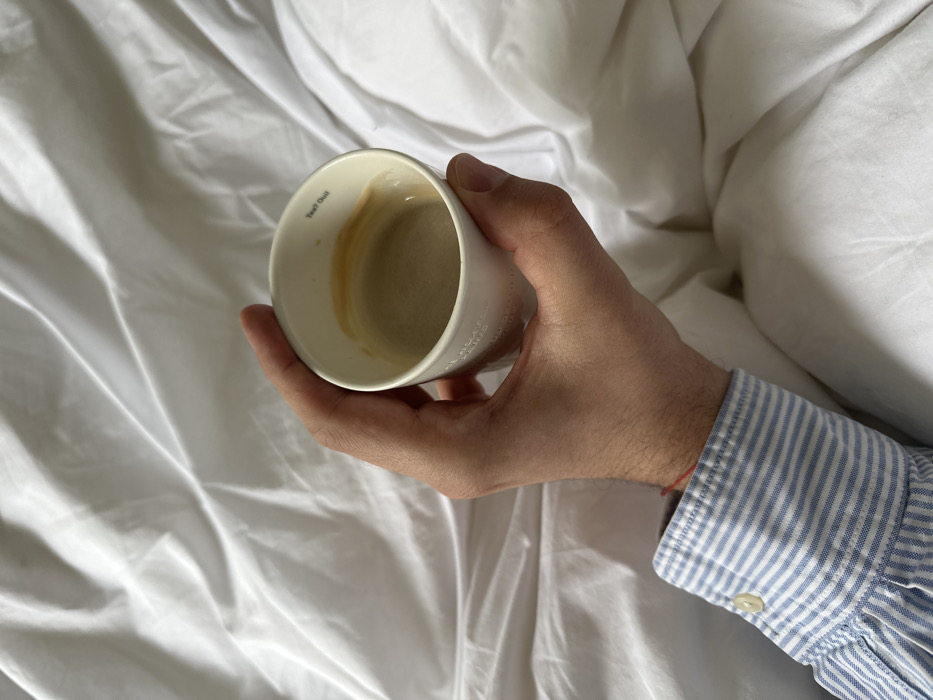 The image displays a close-up, high-angle view of a person's right forearm and hand. The subject, wearing a blue and white striped long-sleeved shirt with a visible buttoned cuff and a thin red string on their wrist, holds a white ceramic cup. The cup contains a residual light brown liquid, consistent with coffee or espresso, with some crema residue on the interior. Text visible inside the cup's rim states "TEA? OUI!", while the exterior bears the partial inscription "THE HOXTON, HOLBORN". The background consists of white, wrinkled bedding, suggesting a hotel room setting. The discernible text on the cup identifies the location as The Hoxton hotel in Holborn, London, United Kingdom.