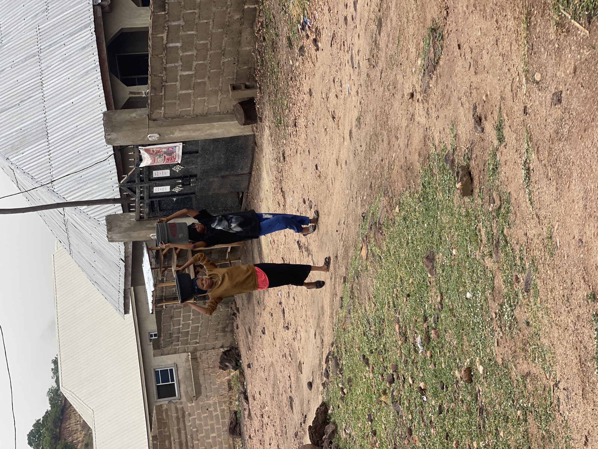 Two individuals are carrying buckets on their heads while standing on a dirt road in Lokoja, Nigeria. The person in the foreground is wearing a brown shirt and black skirt, while the person in the background is wearing a black shirt and blue pants. They are both facing the camera and appear to be smiling. A building with a corrugated metal roof is on the left, and grassy vegetation is on the right. The road is dirt, with some scattered rocks and vegetation. The sky is overcast. The houses nearby appear to be constructed of cinder blocks and concrete.