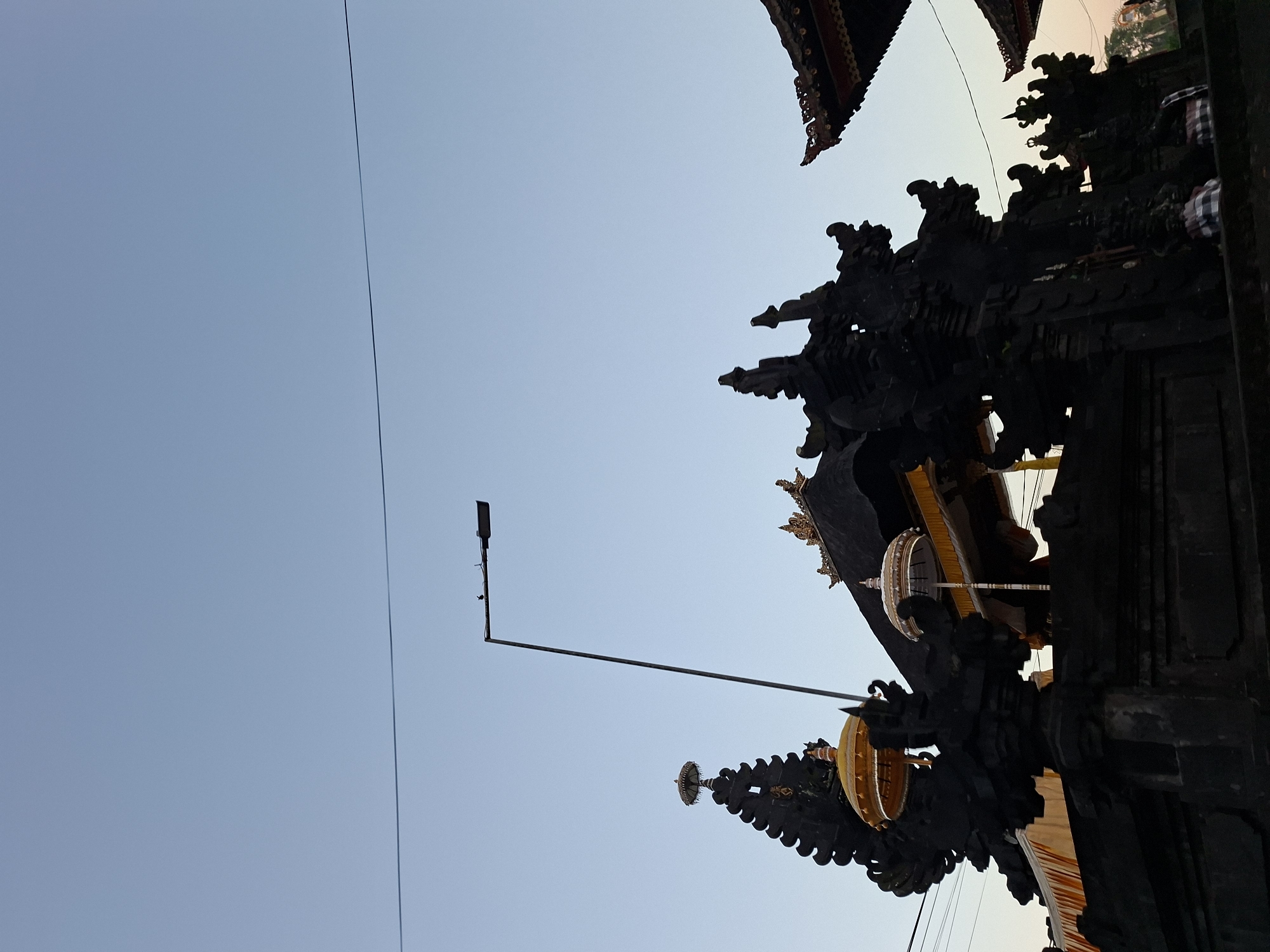 The image depicts the facade of a traditional Balinese temple in Sukawati, Indonesia, under a clear sky at dusk. The foreground is dominated by dark, intricately carved stone structures, characteristic of Balinese architecture, with ornate spires and decorative elements. Two ceremonial umbrellas, one golden and one white, are visible, adding a touch of vibrancy. A street lamp with two small birds perched on it stands tall against the sky, with a thin wire running horizontally across the frame. In the background, hints of other temple roofs with intricate carvings and golden ornamentation are visible. The overall atmosphere is serene, capturing a moment of stillness before the night.