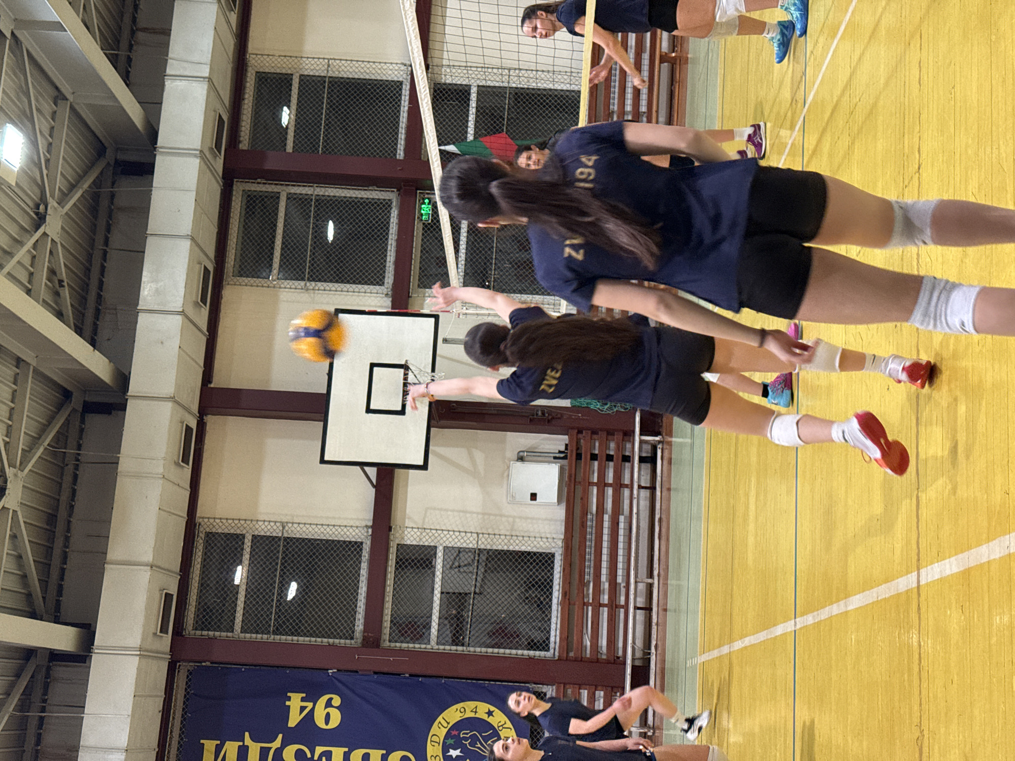 The image shows a volleyball game in progress indoors. The players are wearing dark blue jerseys with black shorts. In the foreground, one player is reaching to hit the ball. The court is yellow. In the background, there are windows with wire mesh. A banner with "ВЕСАМ '94" is visible. The location appears to be in Sofia, Bulgaria, based on the club name displayed on the banner. A basketball hoop is visible on the wall.