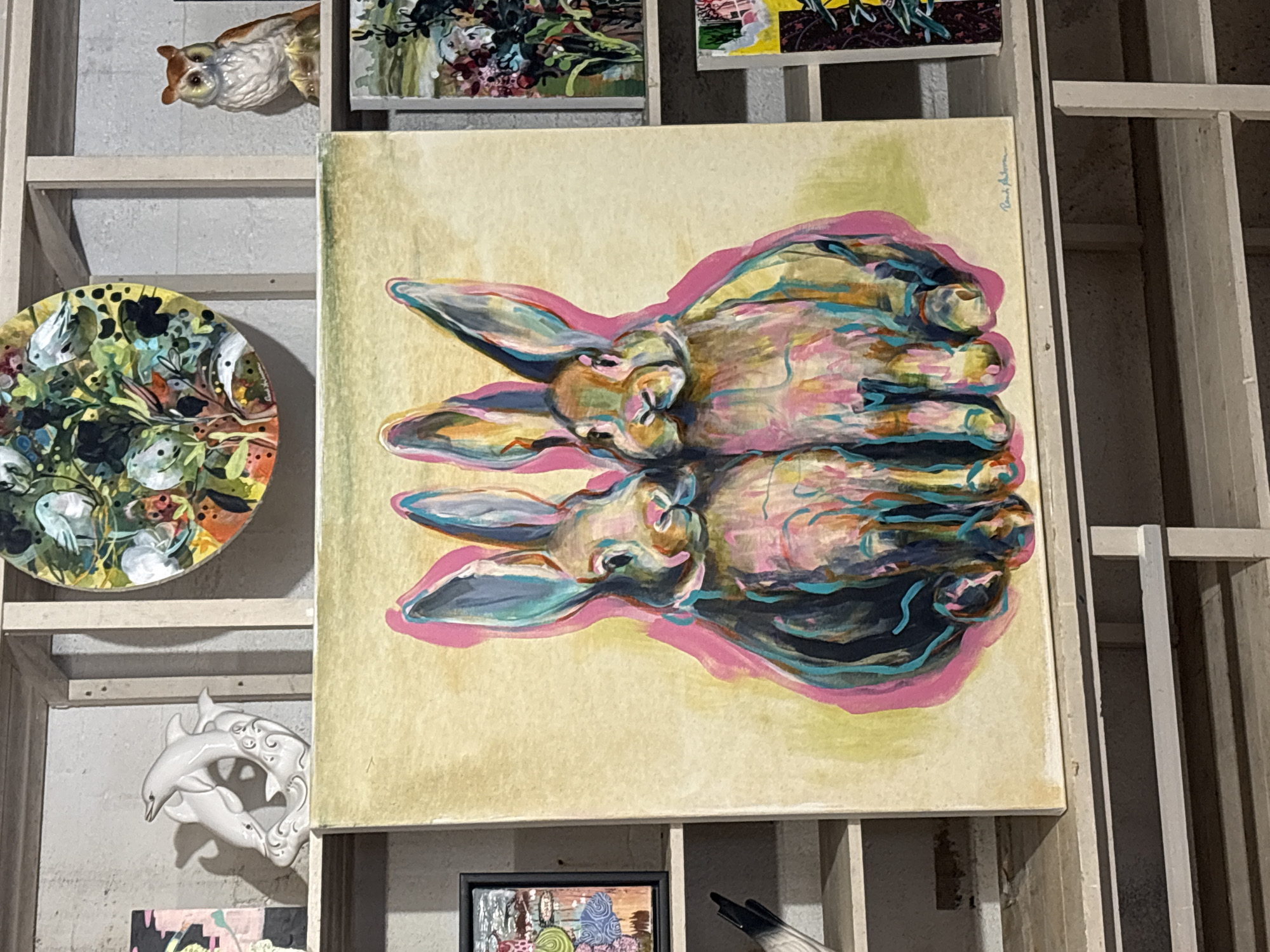 The image shows a painting of two rabbits with long ears, rendered in a colorful and abstract style. The rabbits are depicted sitting side-by-side, with vibrant pink and turquoise hues defining their forms against a muted yellow background. The painting appears to be on canvas and is placed on a shelf, suggesting it is part of a display or collection of artwork.
Above the rabbit painting, on a higher shelf, is a circular piece of art featuring various plants, flowers, and birds. To the right of this circular artwork is a small, ceramic owl figurine with brown and white plumage. To the left of the circular artwork is a white ceramic sculpture of two dolphins leaping.
Further to the left of the rabbit painting, on a shelf below, is a framed artwork with a black border, displaying a collage of colorful shapes and patterns. Below this, on another shelf, is the tip of a dark object, possibly a pen or art tool.
To the right of the rabbit painting, on the same shelf, are two more framed artworks, one with a dark frame and colorful floral imagery, and another below it with a white frame and abstract yellow and dark green elements.
The overall environment appears to be an indoor setting, possibly an art studio, gallery, or storage area, with multiple shelves holding various artworks and decorative objects. The shelves themselves are constructed from light-colored wood or material and are set against a grey, textured wall. The lighting is diffuse, casting soft shadows. There are no people visible in the image.
The prompt requests the location in the city of Gryllefjorden, Norway. However, there are no visual cues within the image that provide any specific information about its geographical location. The scene is consistent with a generic interior space for displaying art and does not contain any identifiable landmarks or cultural markers of Gryllefjorden.