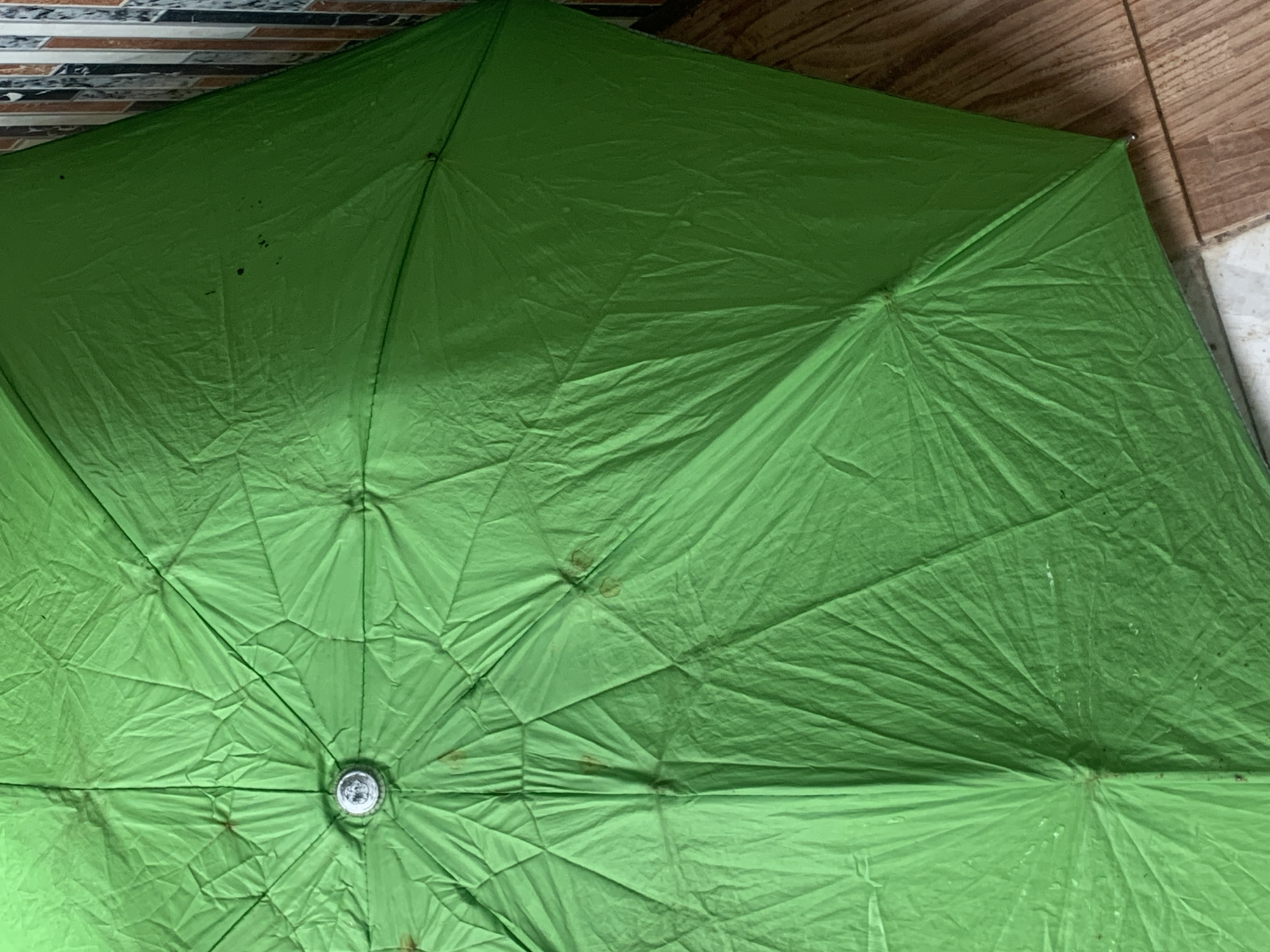 The image shows the top of an open, bright green umbrella with visible creases and some dark spots on its fabric. The center of the umbrella has a metallic button or fastener. The umbrella is lying on a wooden floor, and a tiled wall is partially visible in the background. The scene is indoors or in a sheltered outdoor area, as indicated by the flooring and wall. There are no people or actions depicted. This scene is consistent with an everyday object being stored or displayed. This image was taken in Jauro Yinu, Nigeria.