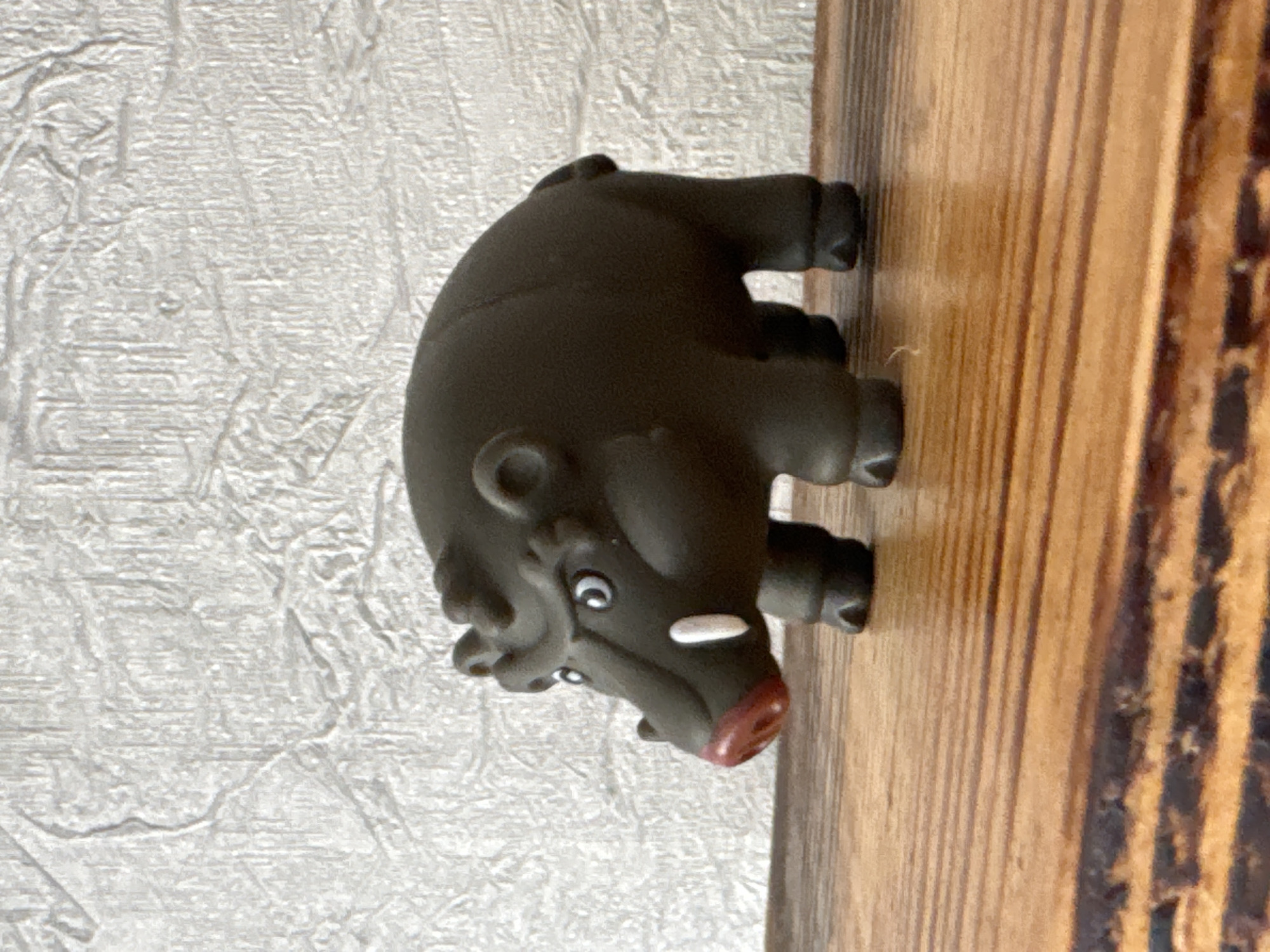 A dark brown toy boar with white tusks and a red nose stands on a wooden surface. The boar is facing left, with its eyes looking forward. Its ears are small and round, and it has a scowl on its face. The background is a textured white wall. The wooden surface appears to be a table or shelf. The scene is lit by natural light. The location of this image is unknown and cannot be determined to be in Яромінскі сельскі Савет, Belarus, as there is no visual information to support this claim.