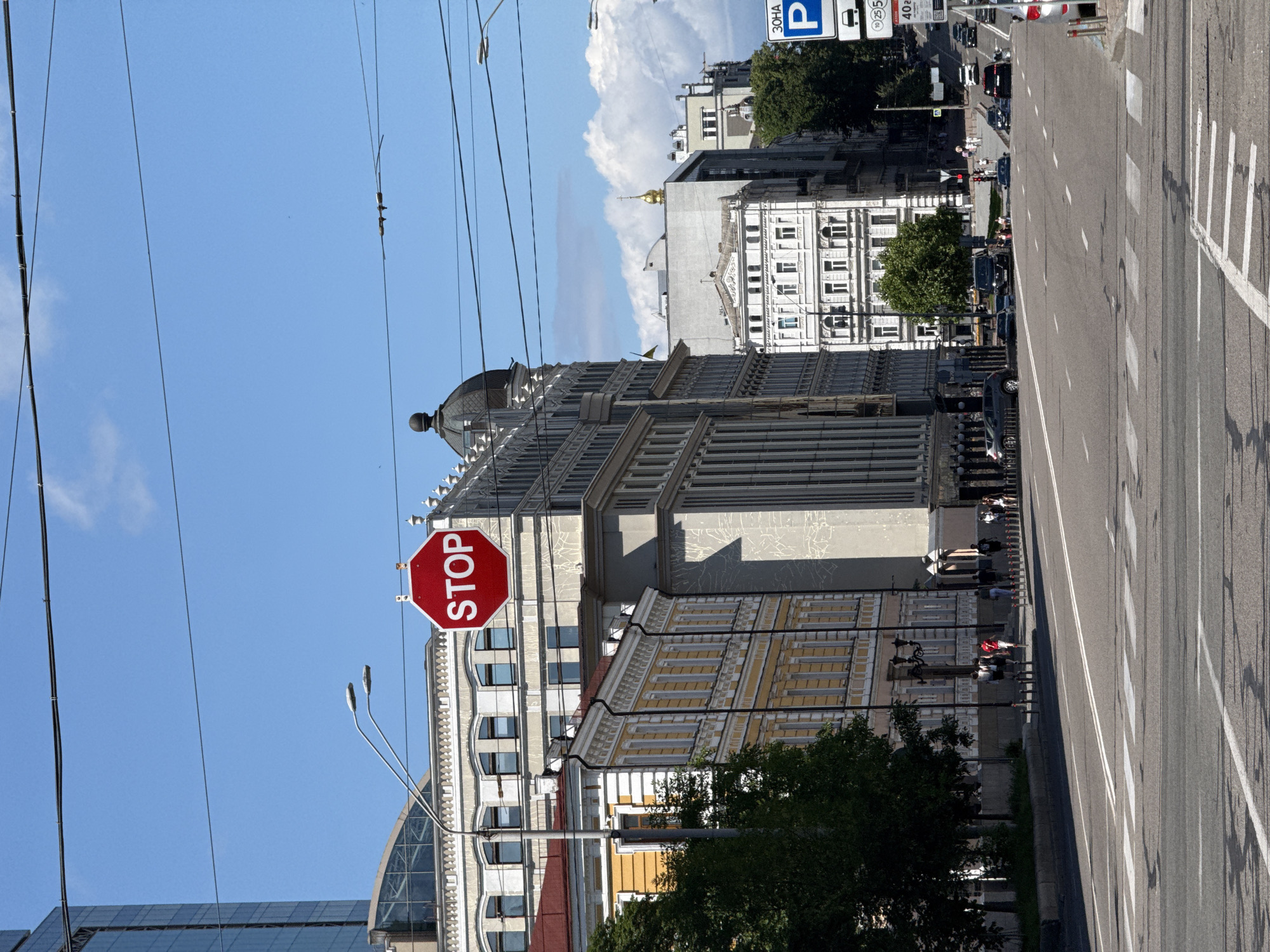 The image depicts a sunny day on a wide city street in Kyiv, Ukraine, characterized by a mix of architectural styles and urban elements. In the foreground, a pedestrian crossing is marked on the asphalt road, with double white lines indicating lanes of traffic. Several cars are visible driving on the road, including a silver sedan parked on the right side of the street.
Dominating the background are multi-story buildings lining the street. On the left, a pale yellow building with ornate detailing and a red roof stands next to a modern, tall glass building. In the center, a prominent octagonal red stop sign hangs from a street lamp pole, clearly visible against the blue sky. Adjacent to it is a light gray building with a rhythmic pattern of windows and decorative elements. Further down the street, more buildings of varying designs can be seen, including one with a golden dome, suggesting the presence of religious or historic structures.
A few pedestrians are walking along the sidewalk on the left side of the street, some in small groups. Trees provide greenery along the sidewalk, interspersed with streetlights and lampposts. Overhead, electrical wires crisscross the sky, typical of an urban environment. Traffic signs, including a parking sign with Ukrainian lettering, are present on the right side of the road. The sky is bright blue with scattered white clouds. The overall impression is a bustling urban scene in Kyiv on a clear day.