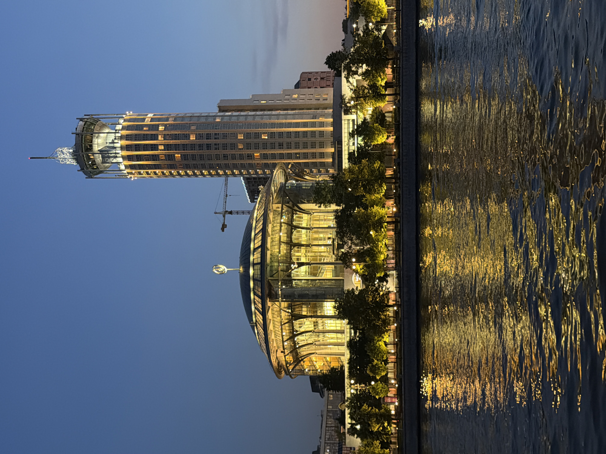 The image depicts a nighttime view of the Moscow International House of Music (MIHM) and the nearby hotel Ukraine, also known as the Radisson Collection Hotel, Moscow. The MIHM, a prominent cultural institution, is characterized by its distinctive domed structure illuminated with warm yellow light, revealing its transparent glass facade and intricate architectural supports. The hotel Ukraine, a Stalinist skyscraper, stands tall to the right of the MIHM, its cylindrical form also lit up with vertical streaks of light, highlighting its numerous windows. Both structures are situated along the Moskva River, whose dark waters reflect the golden glow of the buildings and the surrounding lights, creating a shimmering effect on the surface. Along the riverbank, a row of trees, also illuminated, adds to the picturesque scene. A construction crane is visible in the background, suggesting ongoing development in the area. The sky is a deep twilight blue, indicating that the photograph was taken shortly after sunset. There are no people visible in the image.