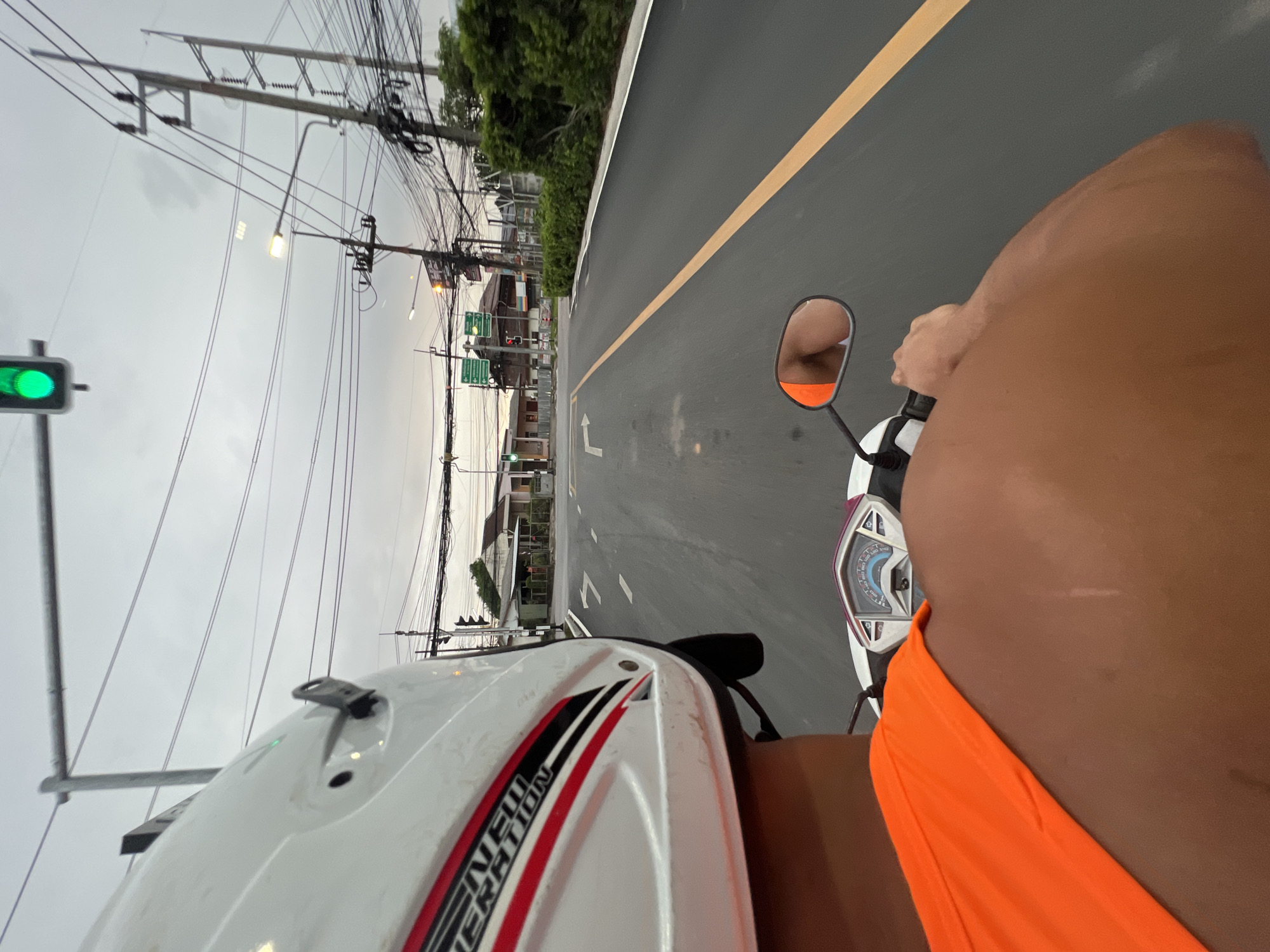 The image was taken from the perspective of someone riding a scooter in Chalong, Thailand. The rider is wearing a white helmet with red and black accents. The rider's bright orange shirt is visible in the foreground. The scooter's dashboard is partially visible. The road ahead shows lane markings and a yellow dividing line. In the background, there are buildings, power lines, streetlights, and a traffic light showing green. The sky is overcast.