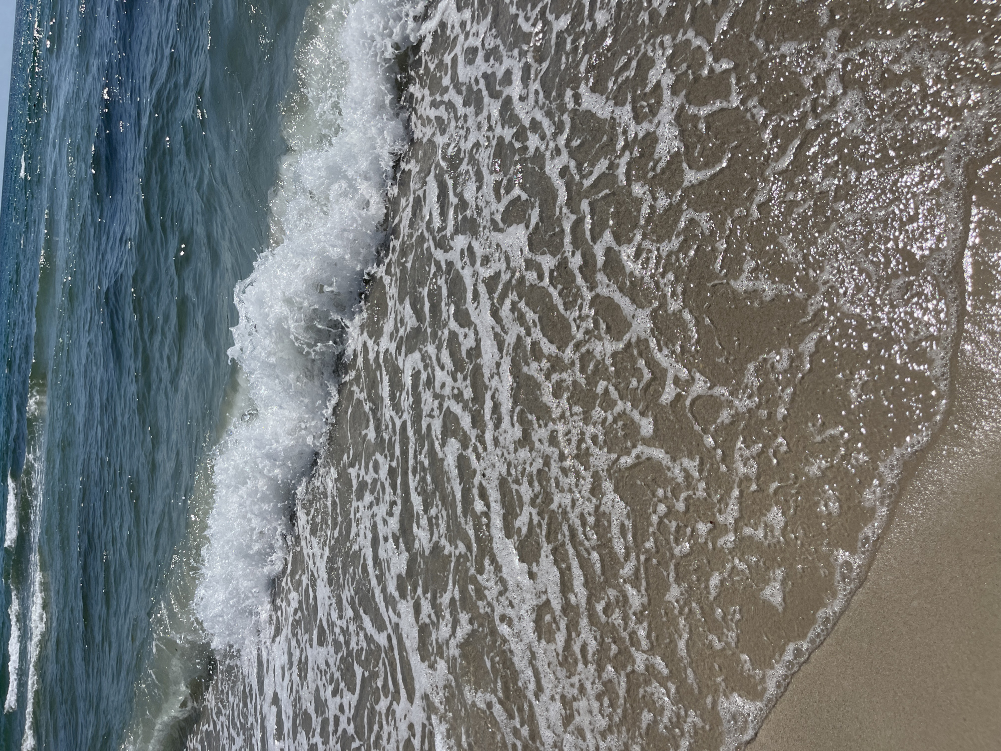 The image captures a close-up, eye-level view of a sandy beach meeting the ocean on a sunny day. In the foreground, wet sand is visible, transitioning to the gentle lapping of shallow waves and foam. The water itself is a deep teal color, with sunlight glinting off its surface, indicating calm conditions. Further out, a series of small waves are breaking, creating white foam as they roll towards the shore. The horizon is visible in the distance, showing a clear blue sky. The scene is devoid of people or any man-made structures, focusing solely on the natural elements of the beach and ocean.