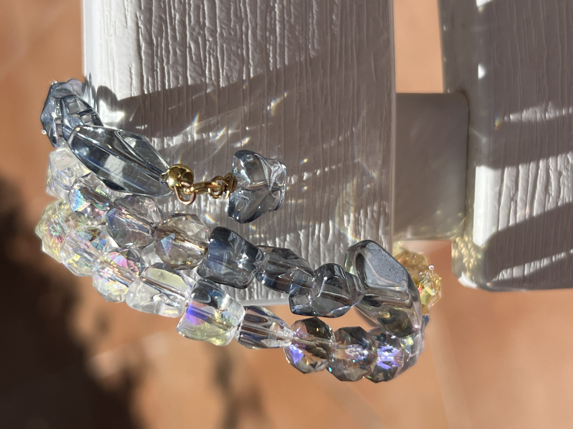 In Bijela, Montenegro, a close-up, shallow depth of field shot shows a bracelet made of faceted, clear, iridescent, and gray beads. The bracelet is draped over a white textured surface, possibly a piece of furniture or railing. A small, gold-colored clasp is visible, with a faceted, light blue heart-shaped charm hanging from it. The background is blurred, with warm, orange-toned lighting, suggesting an outdoor setting, possibly illuminated by sunlight. The lighting creates numerous small, colorful refractions and reflections on the white surface and the bracelet itself. No people or animals are present in the image.