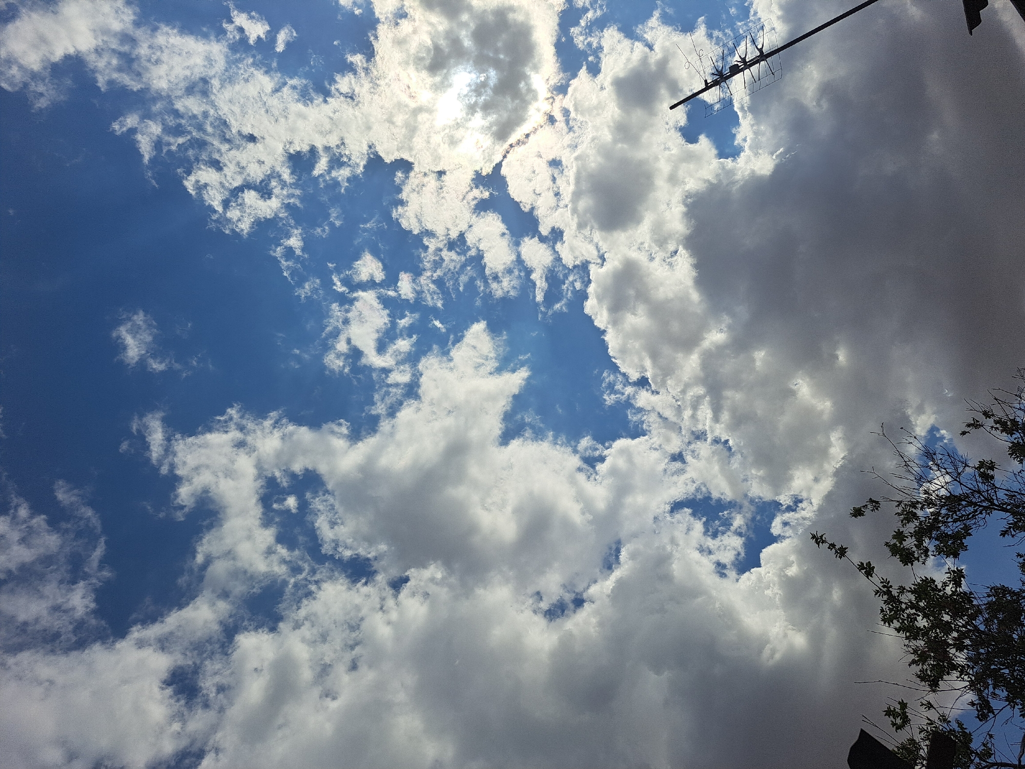 The image captures a dynamic sky with a mix of fluffy white cumulus clouds and darker, more ominous gray clouds against a vibrant blue backdrop. Sunlight filters through the clouds, creating bright highlights and casting shadows. In the lower portion of the frame, silhouetted tree branches and foliage are visible, suggesting an outdoor scene. A television antenna extends diagonally into the sky from the right side of the image. The scene appears to be a clear day with scattered cloud cover, possibly in the city of Vypasne, Ukraine, given the contextual information. No people or specific actions are depicted.
