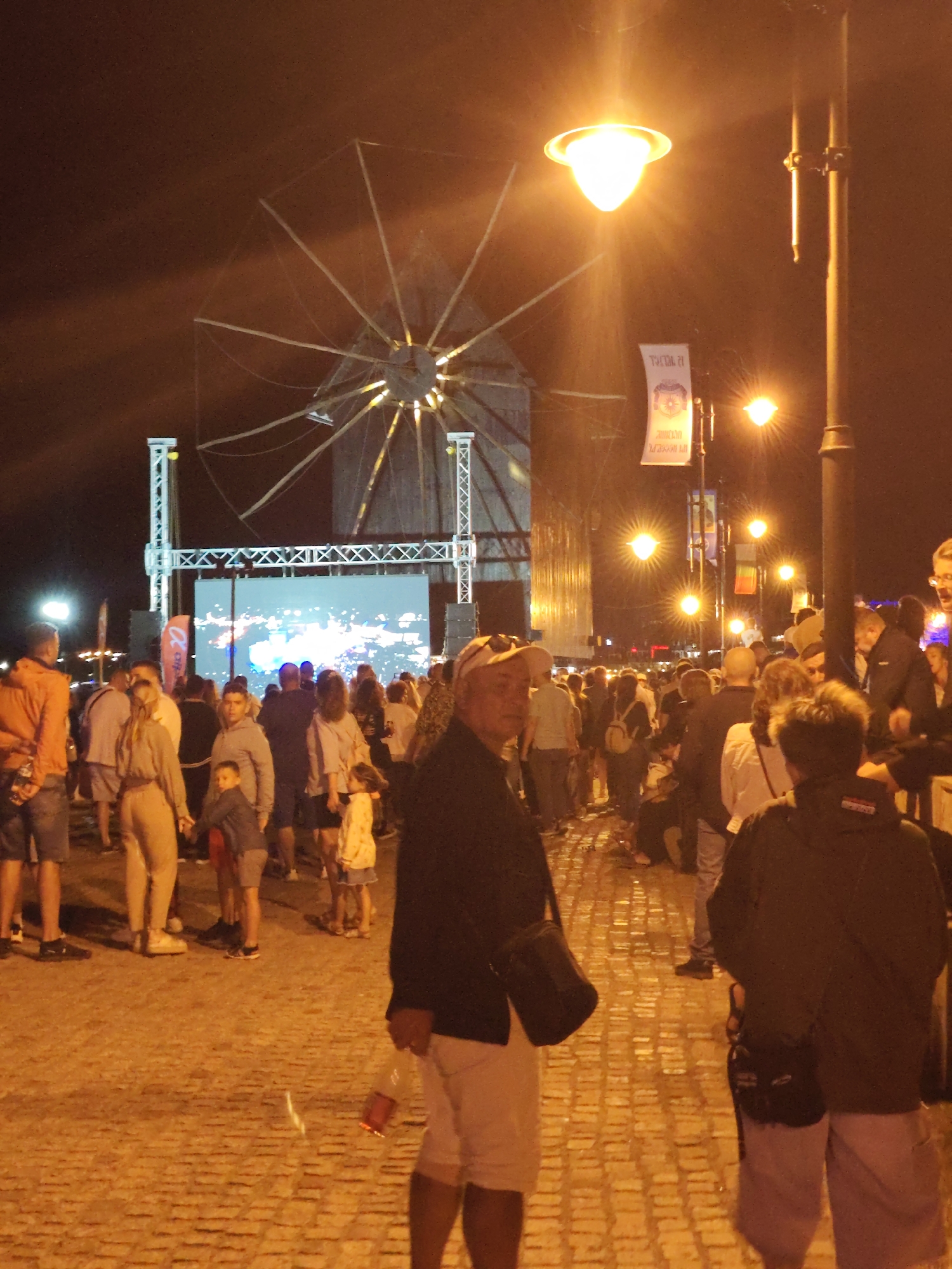 This image depicts a nighttime scene in the city of Nesebar, Bulgaria, likely during a festival or event. In the background, a large, illuminated windmill structure is visible, its arms extending outwards against the dark sky. A large screen or stage is set up in front of the windmill, displaying a bright, abstract light show. Several streetlights illuminate the scene, casting a warm glow on the cobblestone ground and the crowd of people gathered.
The foreground is populated with a diverse group of individuals of varying ages, many of whom appear to be looking towards the stage or interacting with each other. Some are holding drinks, and there is a general atmosphere of a public gathering. Clothing ranges from casual wear like t-shirts and shorts to jackets and hats, appropriate for an outdoor evening event. The lighting creates a somewhat blurred and hazy effect, typical of nighttime photography in an illuminated outdoor setting. The cobblestone ground suggests the location is an older part of the city, consistent with Nesebar's historic architecture.
