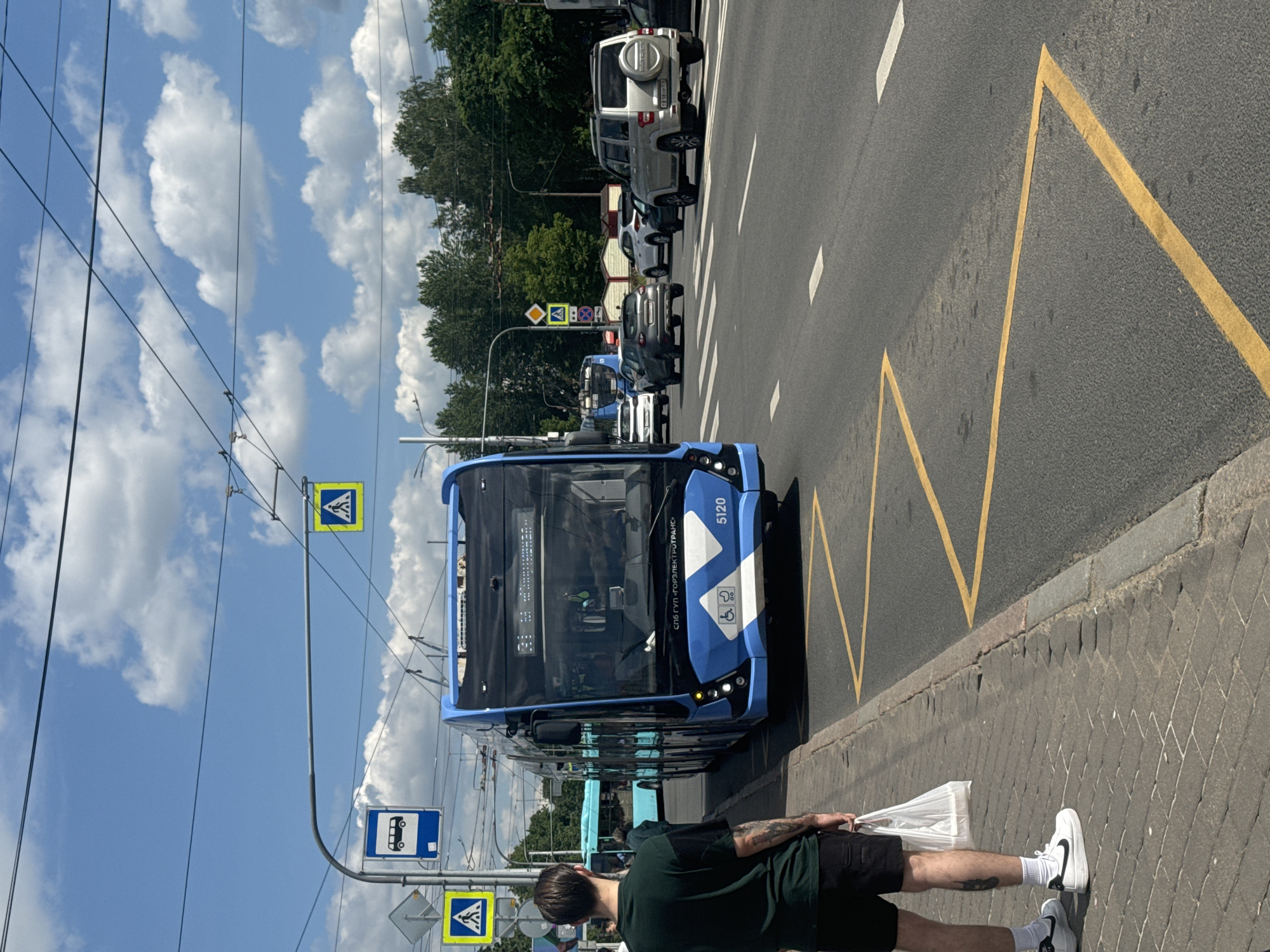 A blue trolleybus with the number 5120 is stopped at a bus stop on a sunny day in Saint Petersburg, Russia. The trolleybus has "СПб ГУП «Горэлектротранс»" written on its front. The bus stop is located on a paved sidewalk next to a road with yellow zig-zag markings indicating a no-parking zone. A person wearing a green t-shirt and black shorts is standing on the sidewalk, holding a white plastic bag. Other vehicles, including cars and another trolleybus, are visible on the road. Overhead power lines for the trolleybus system crisscross the sky, which is blue with scattered white clouds. Traffic signs, including pedestrian crossing and bus stop signs, are present near the bus stop. The environment appears to be a typical urban street scene.