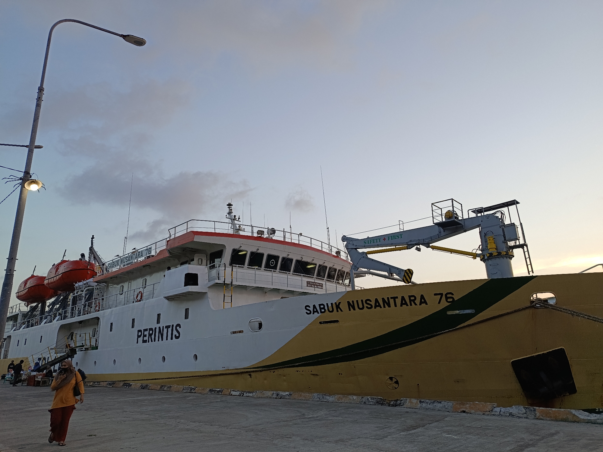 An exterior, low-angle, medium shot captures a large white and yellow ship docked at a pier in the city of Mansalean, Indonesia. The ship, labeled "PERINTIS" and "SABUK NUSANTARA 76", has two orange life rafts mounted on its side. A crane with "SAFETY FIRST" written on it extends from the ship. A person wearing an orange shirt and maroon pants walks away from the camera in the foreground, facing a light pole with a spider-like attachment. Other people are visible in the background near the ship. The sky is a pale blue with scattered clouds.