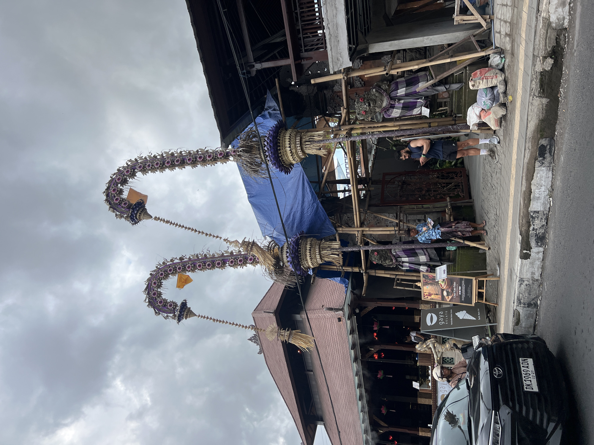 The photograph captures a street scene in Ubud District, Indonesia. The sky overhead is overcast. On the right side of the frame, a black Toyota car is parked, with its license plate visible and reading "DK 1969 ADN." A person on a scooter is situated next to the car.
The focal point of the image features two elaborate, curved floral decorations, each topped with woven baskets and streamers. These decorations seem to be affixed to a building under construction or renovation, indicated by the presence of bamboo scaffolding and a blue tarp covering a section of the structure. The building has a red-tiled roof.
A man, clad in a black tank top and shorts, stands on the sidewalk with his head down, apparently using his phone. Several signs indicating a business establishment, possibly a restaurant or café named "Gaya Gelato," are visible. Additional decorations and wrapped items are positioned near the scaffolding. The scene gives the impression of ongoing activity amidst traditional Balinese ornamentation.