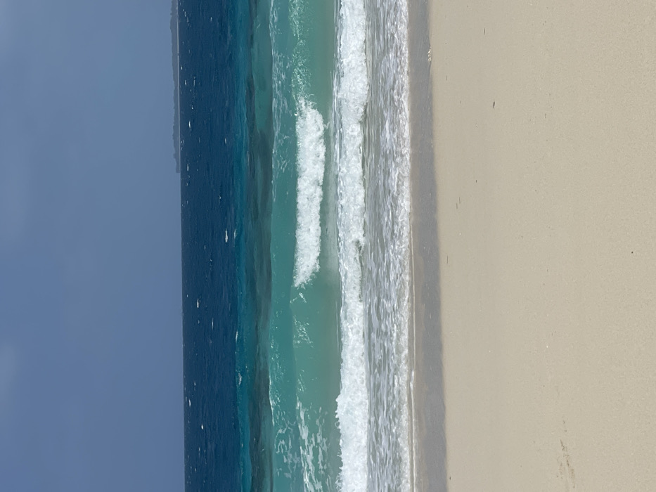 The image depicts a tropical coastal scene on the island of Zanzibar, Tanzania. The foreground consists of smooth, light beige sand, wet at the immediate waterline. The midground features the Indian Ocean with multiple wave formations. Closest to the viewer, a line of white foam from a receding wave extends across the beach. Further out, a larger, clear turquoise wave is cresting, approximately mid-frame. Beyond this, the water transitions to a deeper blue-green, exhibiting surface chop and small whitecaps indicative of wind. The horizon line displays a dark blue ocean meeting a hazy, light blue-gray sky. A faint, low-lying landmass or island is visible on the far right of the distant horizon. There are no identifiable human subjects, animals, or man-made objects within the visible frame.