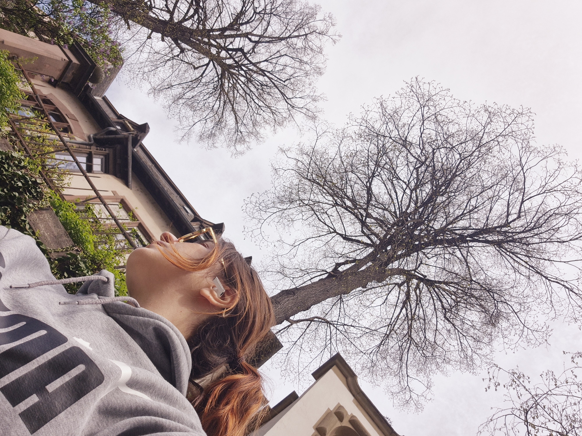 A low-angle shot features a woman looking up at bare trees and buildings in Strasbourg, France. The woman, angled diagonally in the frame, has auburn hair, wears gold-rimmed glasses, and a gray hooded sweatshirt with a large black logo. She appears to be wearing an earpiece in her left ear. Two leafless trees dominate the sky. The building to the left of the woman has several windows and is partially covered in ivy. The building to the right of the woman has a pointed roof and Gothic architecture details. The sky is overcast.