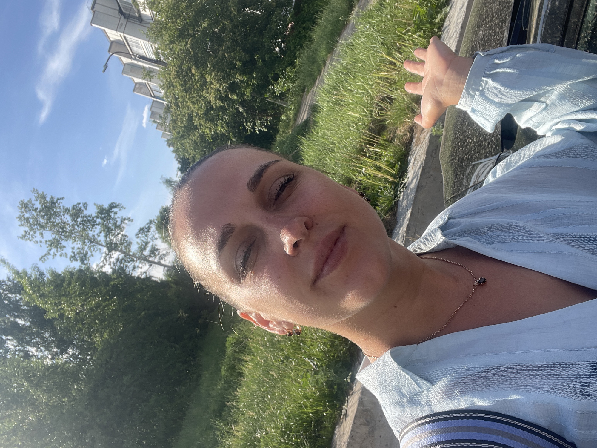 A woman is taking a selfie outdoors. She has fair skin, dark hair pulled back, and is wearing a light blue blouse. She has a gold necklace and earrings. Her eyes are closed slightly, and she is holding her hand up, possibly to shield her face from the sun.
The background shows greenery including grass and trees. In the upper left, a building with white rectangular protrusions is visible against a blue sky with some clouds. The location may be near Chaikovsky, Russia, based on the prompt. There is also what appears to be a car visible behind the woman's hand.