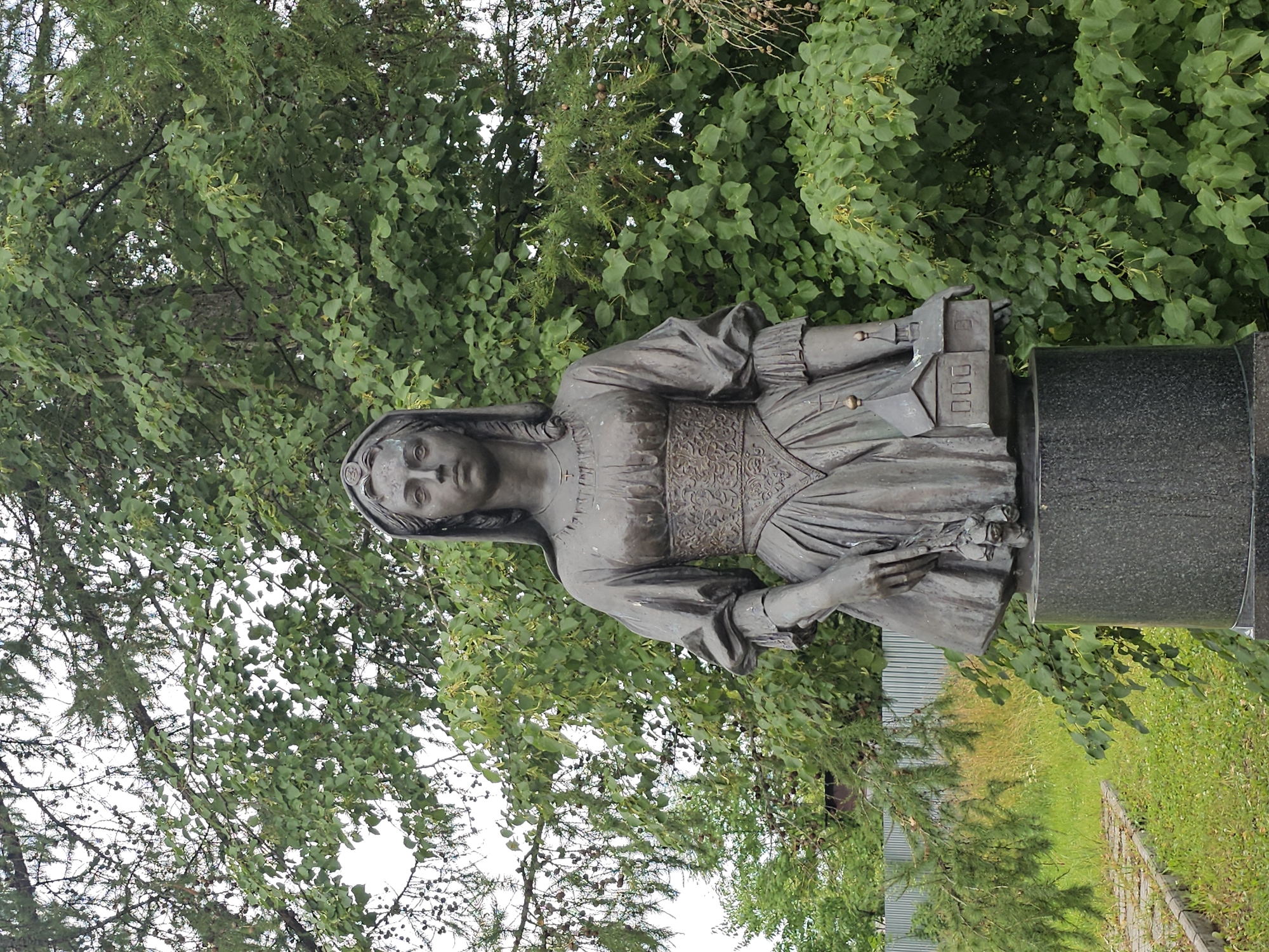 This is a medium shot, eye-level photograph of a bronze statue of a woman in front of lush green trees. The statue depicts a woman in historical attire, wearing a long dress with puffed sleeves and an ornate, patterned bodice. She has a headpiece or veil. In her left hand, she holds a bouquet of roses, and in her right hand, she holds a miniature model of a building with a peaked roof and windows. The statue stands on a dark, granite pedestal. The background consists of dense green foliage, suggesting an outdoor setting, likely a park or garden. There is a glimpse of a gray corrugated metal fence and some dry grass to the left of the statue, indicating it's an outdoor public space. The sky is overcast, with a white, hazy appearance. The statue appears to be located in the city of Ulitkino, Russia, as suggested by the context.