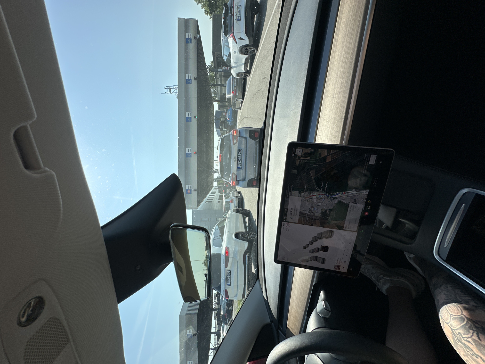 A first-person perspective captures the interior of a Tesla vehicle and the view through its windshield, depicting a queue of vehicles at a multi-lane international border crossing under bright, sunny conditions. The location is identified as Kapitan Andreevo, Bulgaria.
The observer is positioned in the driver's seat of a Tesla, indicated by the steering wheel, wooden dashboard trim, and a prominent central touchscreen display. A tattooed left forearm and hand are visible on the lower left, along with parts of the driver's legs wearing light-colored sneakers, resting on the pedals. The touchscreen displays a map interface with traffic conditions, showing several vehicle icons, and a destination of "22 km." Music playback for "Elton John" is also visible on the screen. An overhead console with a speaker grille and a small sensor/camera is seen above the rearview mirror.
Through the windshield, a line of cars is visible ahead, stationary or moving slowly in multiple lanes leading to border control booths. Directly in the rearview mirror, a white Mercedes-Benz GLS SUV with German license plate "M RQ 580" is visible. In the lanes ahead, a grey Subaru XV/Crosstrek with Bulgarian license plate "CB 4213 XM" occupies the left lane, while a white SUV (possibly a Voyah Free) with Bulgarian license plate "PB 2293 TE" is in the right lane. A dark sedan is partially visible behind the Subaru.
Overhead, a large gantry spans the width of the road, marked with distinct signage for different vehicle types and border procedures. Text on the gantries includes "CARS," "BUSES," and "КАМИОНИ" (Cyrillic for "TRUCKS"), along with other Cyrillic script. Green lights are visible on some of the gantry structures, indicating open lanes. The overall infrastructure suggests a busy and established border crossing. The sky is clear blue with no clouds. Various buildings associated with the border facility are visible in the background, including one with a small antenna tower on its roof.