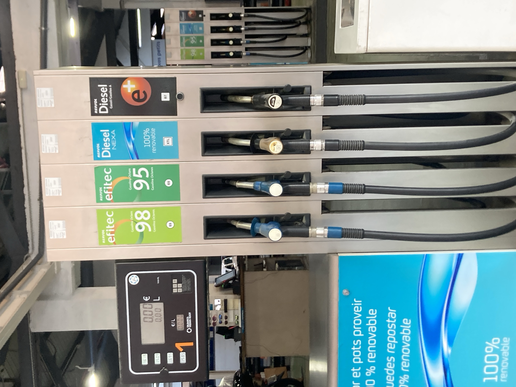 This is an image of a gas station pump in Barcelona, Spain. The scene features multiple fuel pumps with various types of fuel labeled, including "Diesel," "Nexa," and "efitec" gasoline with octane ratings of 95 and 98. The pumps are organized vertically, with hoses and nozzles clearly visible. Below the fuel selection panels, there is a digital display showing a price of "0.00€" and a volume of "0,00 L," indicating no fuel has been dispensed yet. A price per liter of "0.000" is also displayed.
To the right of the fuel pumps, a large blue advertisement is partially visible, with text in Spanish and French that includes "100% renovable." The overall setting appears to be outdoors, with the lighting suggesting it could be daytime. There are no people visible in the image. The focus is on the operational aspects of the gas station, highlighting the different fuel options and the payment display. The image is oriented in a way that the top of the gas station equipment is towards the left side of the frame.