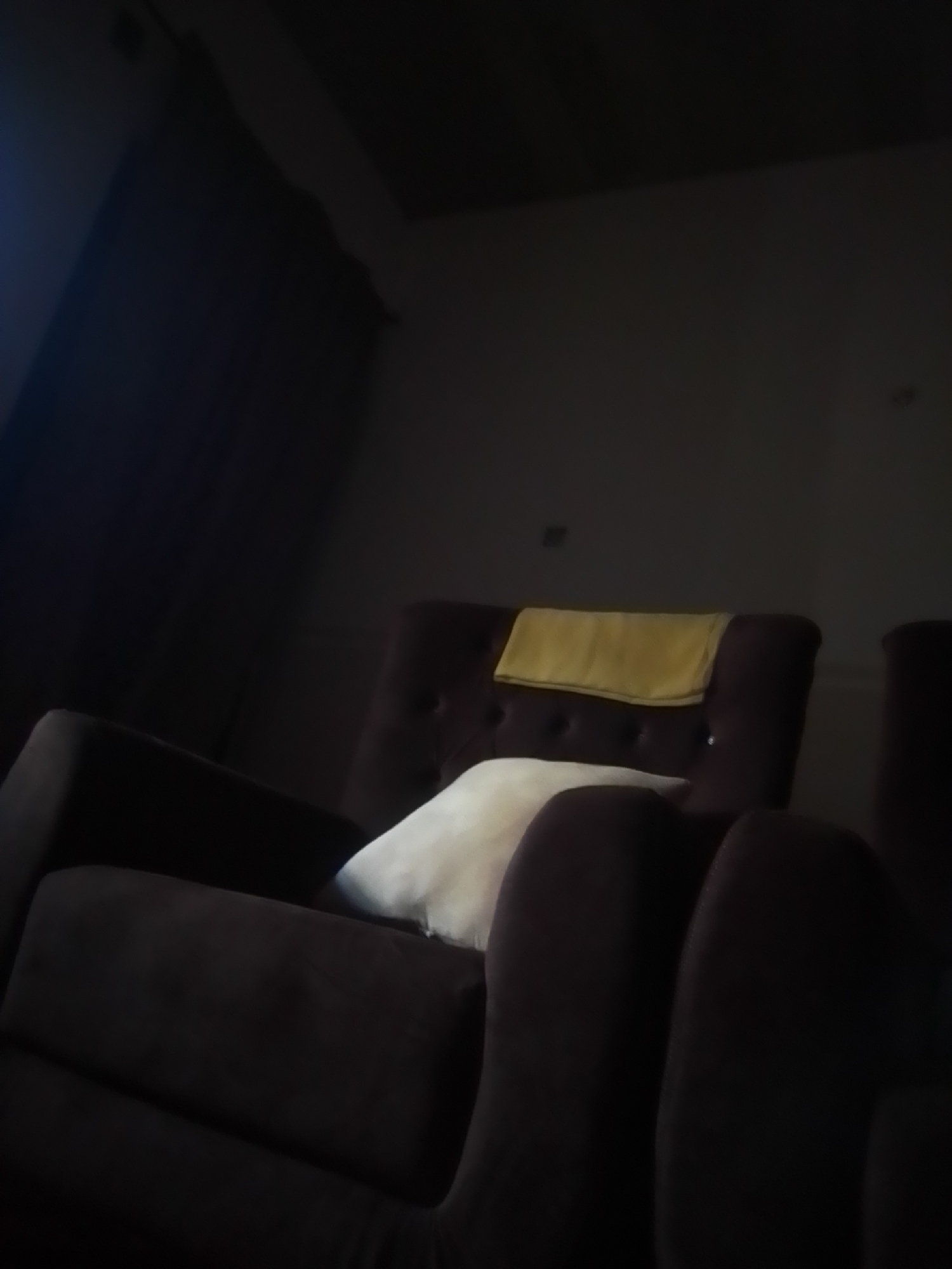 An indoor scene, dimly lit, shows a portion of a dark brown, plush armchair with a tufted back. A white pillow rests on the seat of the armchair, and a folded yellow blanket is draped over the top of the backrest. The background is dark and indistinct, with a hint of a window or curtain on the left side. The scene is likely a living room or lounge area. There are no people visible in the image. The location in the city of Talasse, Nigeria, is not determinable from the visual information provided in the image.
