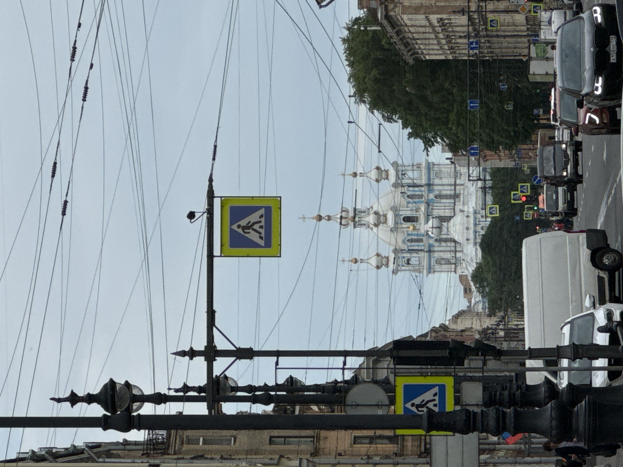 This is a vertical, eye-level, medium shot of a street scene in Saint Petersburg, Russia. The image is dominated by a complex web of overhead wires, common in older cityscapes. In the background, a grand white and blue cathedral with multiple domes and spires stands majestically under a pale, overcast sky, suggesting a cloudy day.
In the foreground, a pedestrian crossing sign with a blue square and white pedestrian symbol is prominently displayed on a traffic signal pole. Another similar sign is visible further down the street. The street itself is lined with buildings in a classical architectural style, some appearing to be in disrepair.
The street is busy with traffic, including several cars and vans. The vehicles appear to be moving slowly, suggesting a traffic jam or a bustling urban environment. The presence of pedestrian crossing signs and traffic lights indicates a functioning road infrastructure within the city.
The overall impression is one of a vibrant, historic city, with elements of both grand architecture and everyday urban life. The overcast sky contributes to a somewhat subdued atmosphere, but the scene is lively with activity. There is no discernible text in the image, other than what might be on vehicle license plates, which are not legible.
