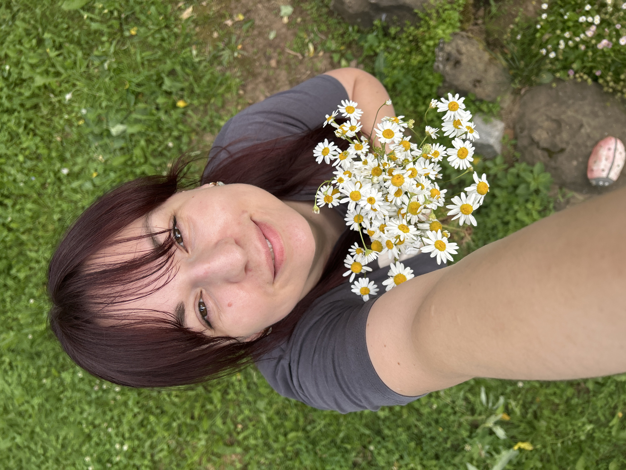 A woman is taking a selfie outdoors with a bouquet of daisies. She has dark hair and is wearing a gray t-shirt. The camera angle is from above, looking down at her face. She is smiling. In the background, there is green grass, some rocks, and a pink ladybug decoration. The scene suggests it is daytime and the weather is likely pleasant, suitable for being outdoors. The woman appears relaxed and happy. There is no visible text in the image. The location context provided is Фоки, Russia.