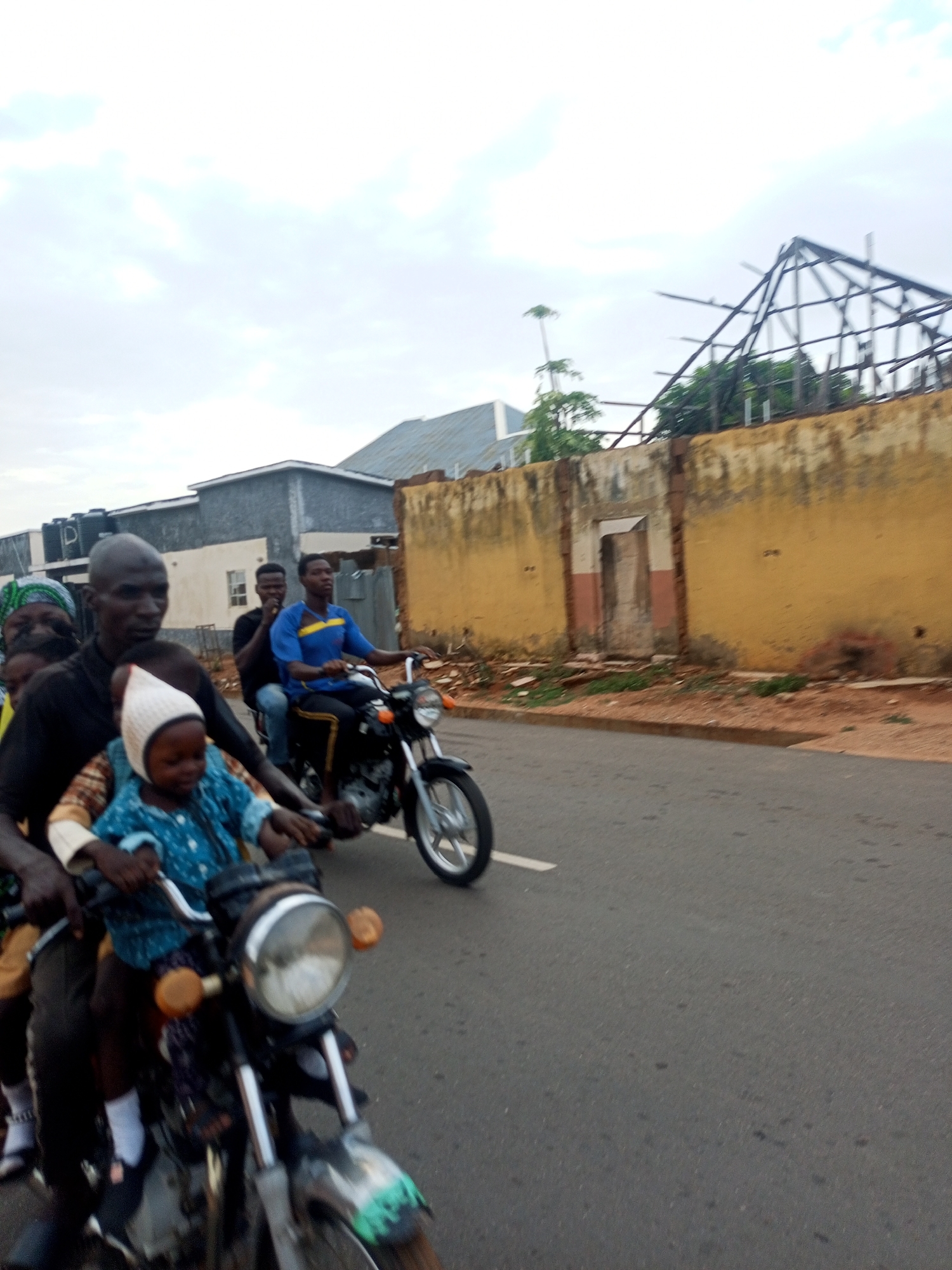 Two motorcycles travel down a street. The first motorcycle, in the foreground, is driven by a dark-skinned man wearing a black shirt and carrying several passengers. At least one passenger in front of the driver is a child in a blue jacket and white hat. The second motorcycle, in the background, is driven by a man wearing a blue shirt, with a man in a black shirt behind him. Buildings, a wall, and exposed metal framing are visible in the background. The scene appears to be set in Tempure, Nigeria.