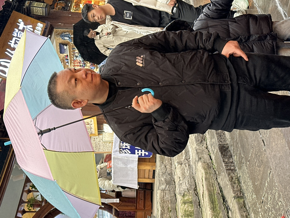 A man holds a colorful umbrella while standing on stone steps. He wears a black jacket and pants. In the background, other people are present, with one wearing a black shirt and another wearing a white jacket. Behind them, a shop or establishment is visible. The umbrella has alternating panels of pastel colors: blue, pink, and yellow. The scene appears to be outdoors, possibly in a market or tourist area.