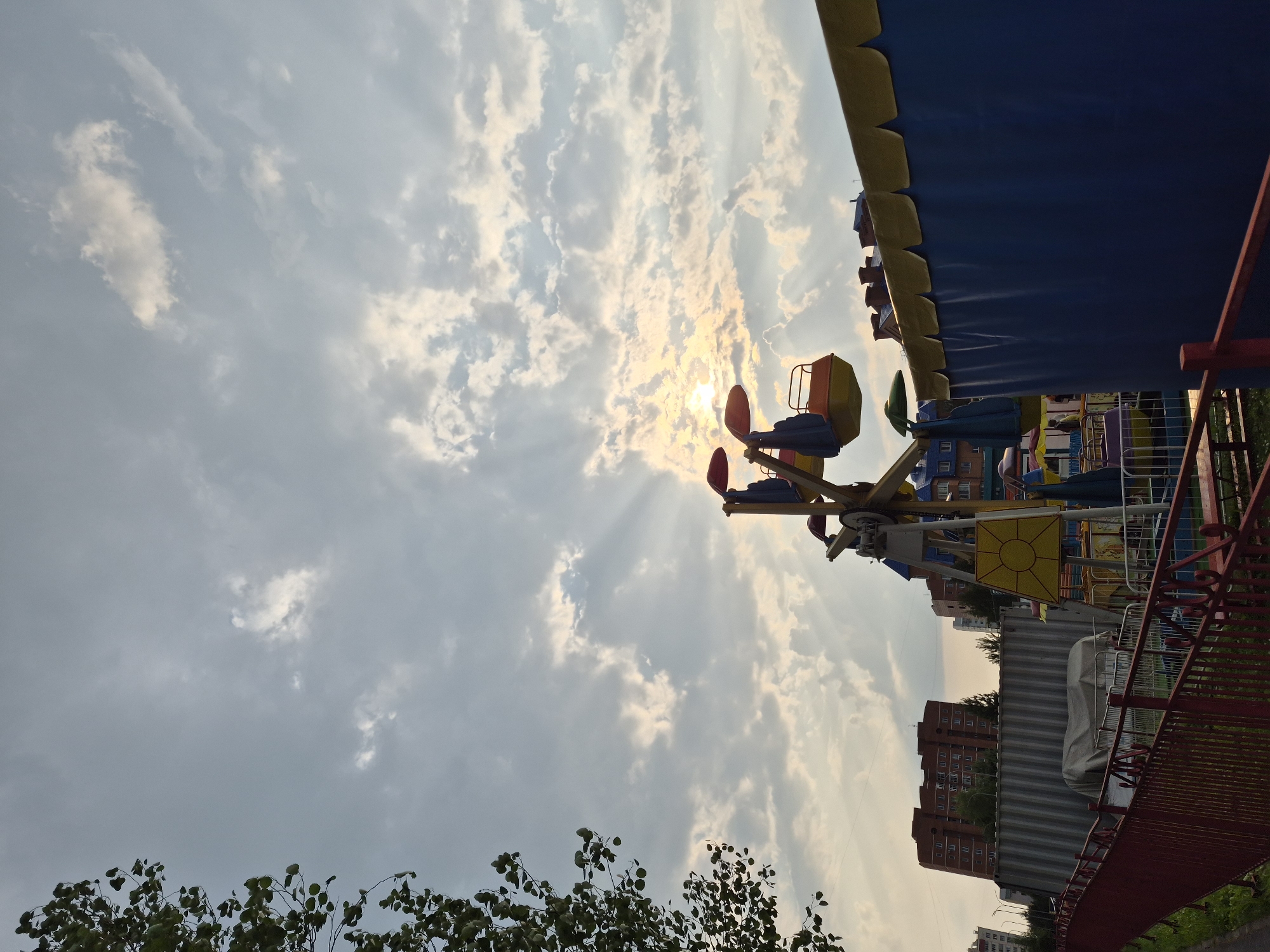 The image shows a view looking upwards at a carnival ride under a cloudy sky with the sun breaking through, creating crepuscular rays. The ride, likely a type of swing ride, has colorful gondolas and is partially obscured by a blue awning with yellow trim. In the background, there are apartment buildings. The presence of the ride and buildings suggests an urban or suburban setting, possibly a park or amusement area. The sunbeams indicate it is likely late afternoon or early evening. The weather appears partly cloudy, with the sun attempting to shine through. There are trees visible in the foreground. No people are clearly discernible in the image. No text is visible. The location context is Nyagan, Russia.