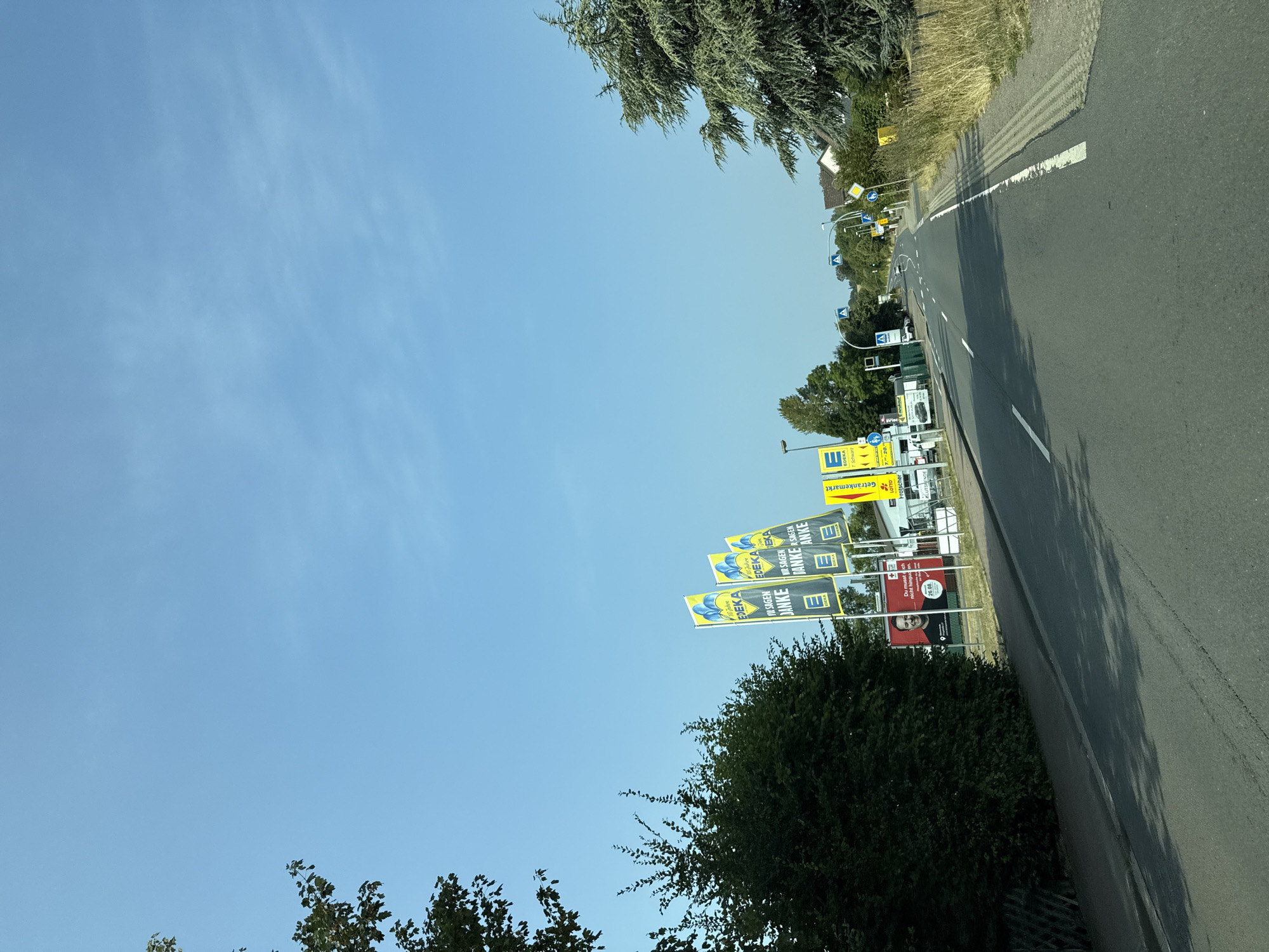 This image depicts a road in the city of Bad Sachsa, Germany. The scene is set on a clear day with a bright blue sky and sparse clouds. On the left side of the road, a dense green hedge lines the shoulder, with a large tree with dark green foliage at the far left edge of the frame. Further along the road on the left, several yellow flags bearing the EDEKA logo and the text "WIR SAGEN DANKE" (We say thank you) are displayed, along with a billboard featuring a smiling man and the text "Du hast mich mutig gemacht. Ich wünsche dir ein schönes Leben." (You made me brave. I wish you a nice life.) and "26.08." (August 26th). Next to the flags, a building with a white facade is visible, with signs indicating "Getränkemarkt" (Beverage market) and other store names.
The road itself is asphalt and curves gently to the right, leading into the distance. There are lane markings on the road, and a white line marking the edge of the road. On the right side of the road, there is a grassy verge with dry vegetation and a tall pine tree. Various road signs, including a yellow diamond-shaped sign with a black arrow and a pedestrian crossing sign, are visible along the road. A few vehicles are present on the road in the distance, including a white car. The overall impression is one of a commercial area or a road leading to amenities, with a focus on a supermarket (EDEKA) and surrounding businesses.