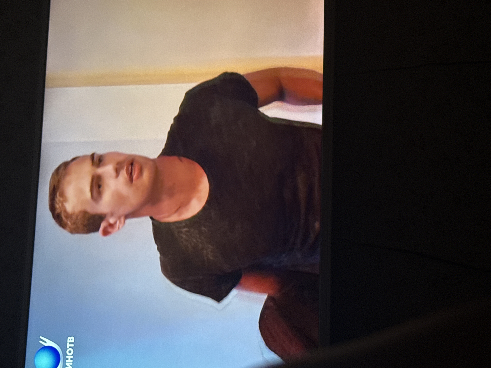 A fair-skinned man with reddish-brown hair and a black t-shirt is on a television screen displaying a blue logo with Cyrillic text "ИНОТВ" in the bottom left corner. The background behind the man appears to be a white wall with a beige trim near the top. The image appears to be taken from an angle looking at the television screen. The man seems to be looking down and to the side. The television itself has a thick black frame. The location in Mytishchi, Russia is unknown.