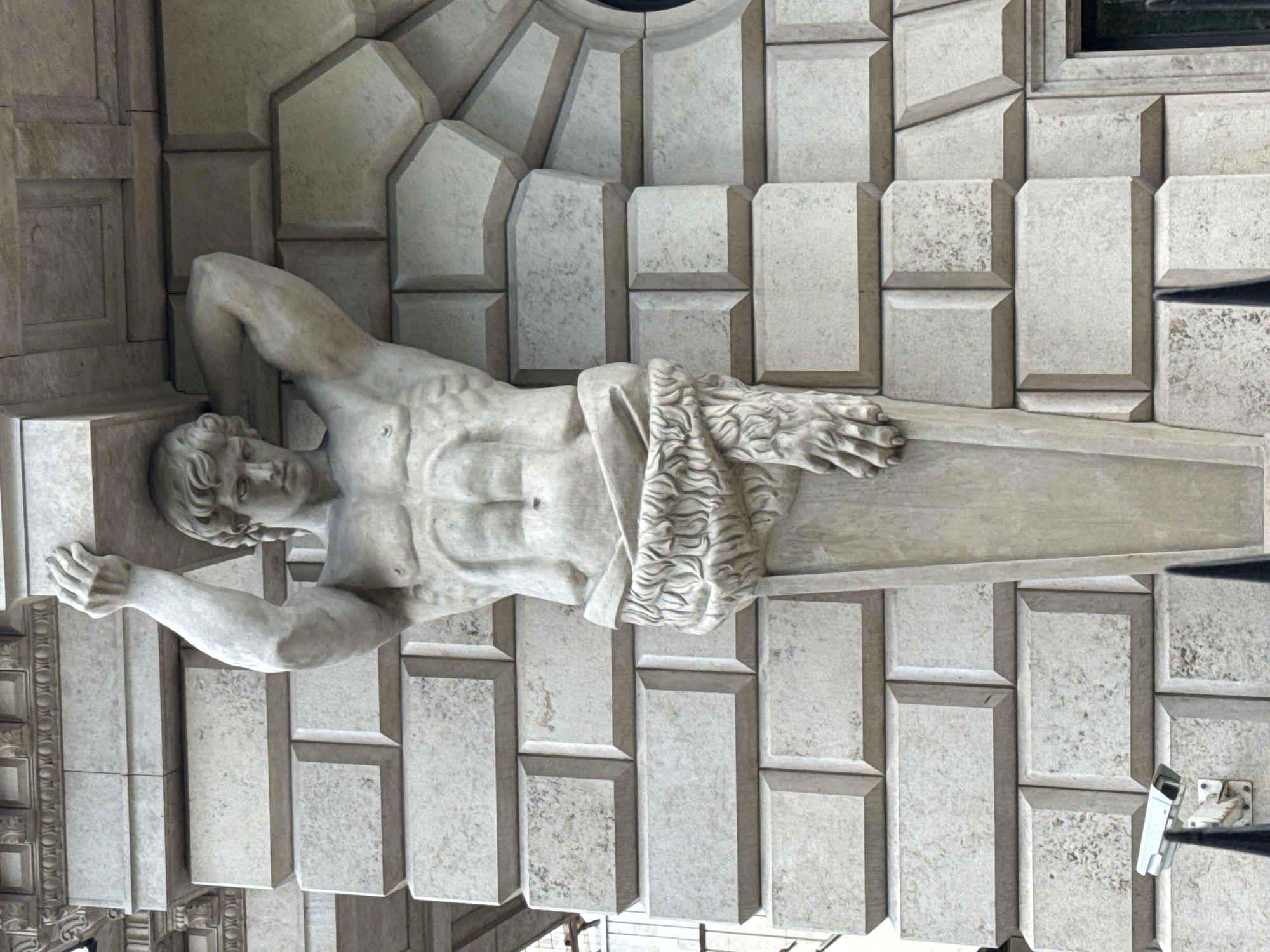 The image shows a horizontal view of a classical architectural detail featuring a sculpted male figure, likely a Telamon, supporting an entablature. The figure is positioned horizontally, with his head resting on a block and his body extending along the structure. A draped cloth or garment covers part of his midsection. The building is constructed with light-colored stone blocks arranged in a regular pattern around the statue. A security camera is visible on the lower right. This architectural detail is located on the facade of the New Hermitage Museum in Saint Petersburg, Russia.