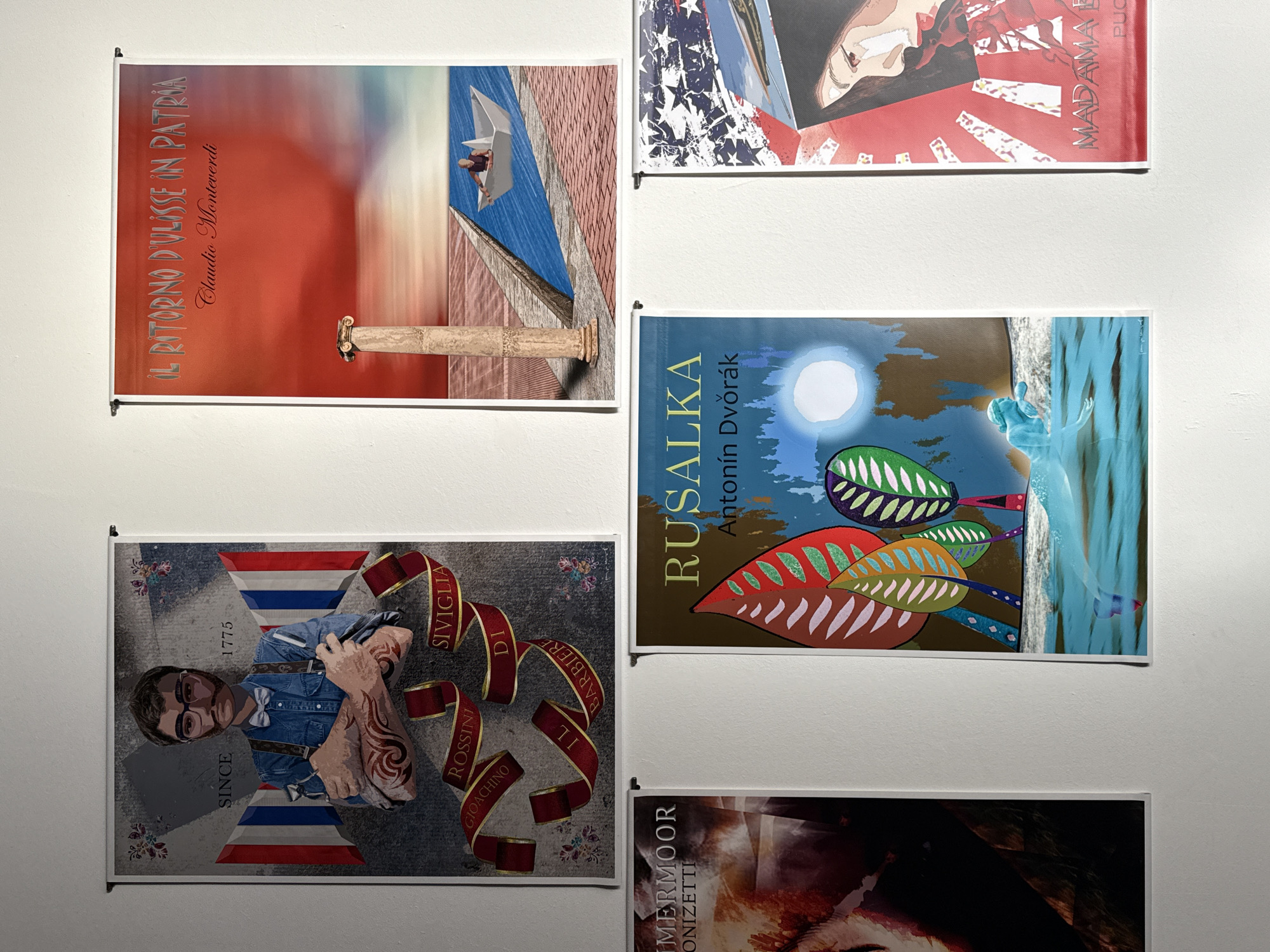 The image depicts a collection of posters hanging on a white wall. These posters seem to be artistic representations or advertisements for various theatrical or musical performances. The subjects vary, including figures in stylized poses, abstract compositions, and thematic designs incorporating text and imagery. The posters are arranged in a grid-like fashion on the wall. The location is not specified, nor are there identifying details to pinpoint it to the city of Barcelona, Spain.